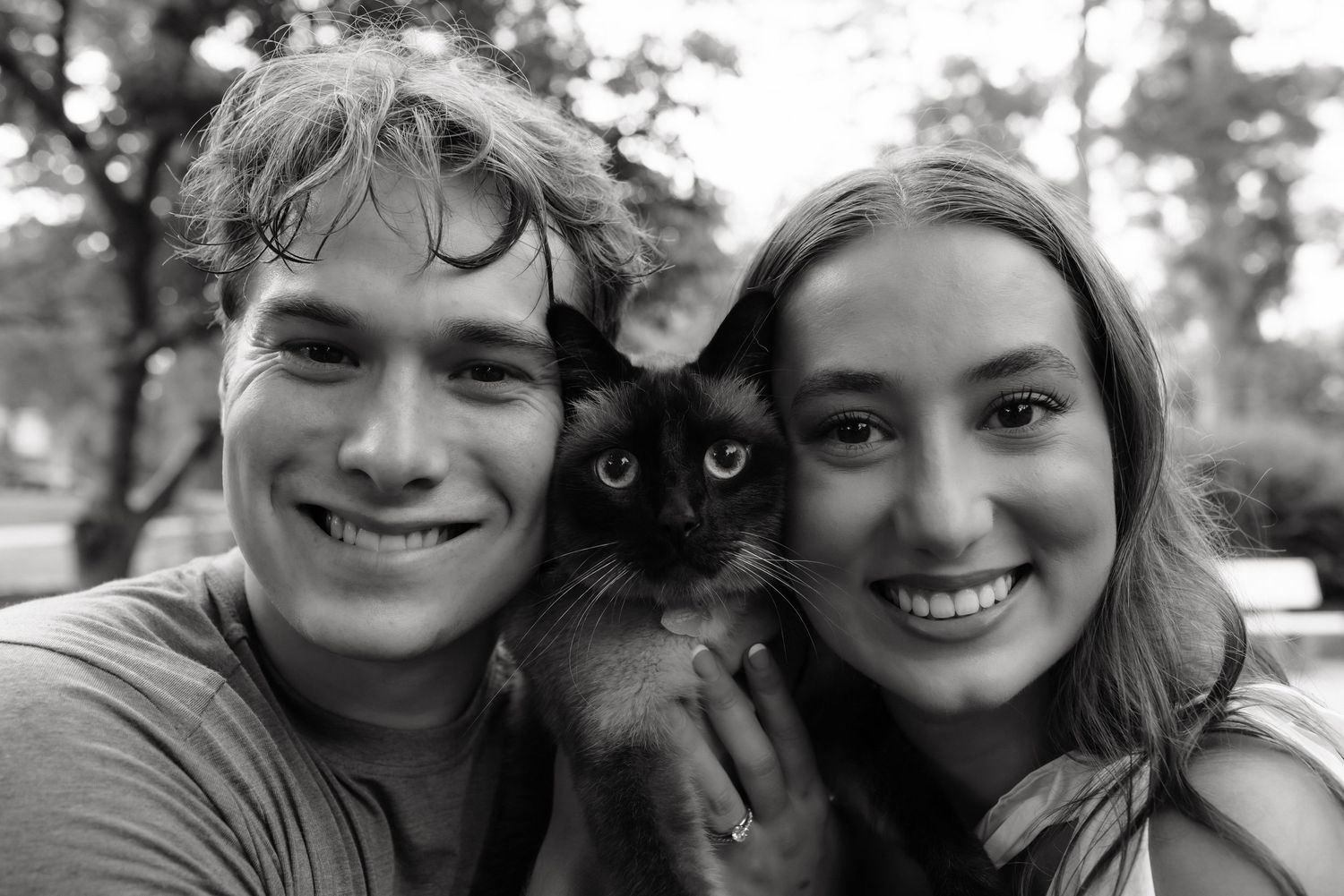 Black and white photo of a smiling couple holding a dark Siamese kitten outdoors among trees.