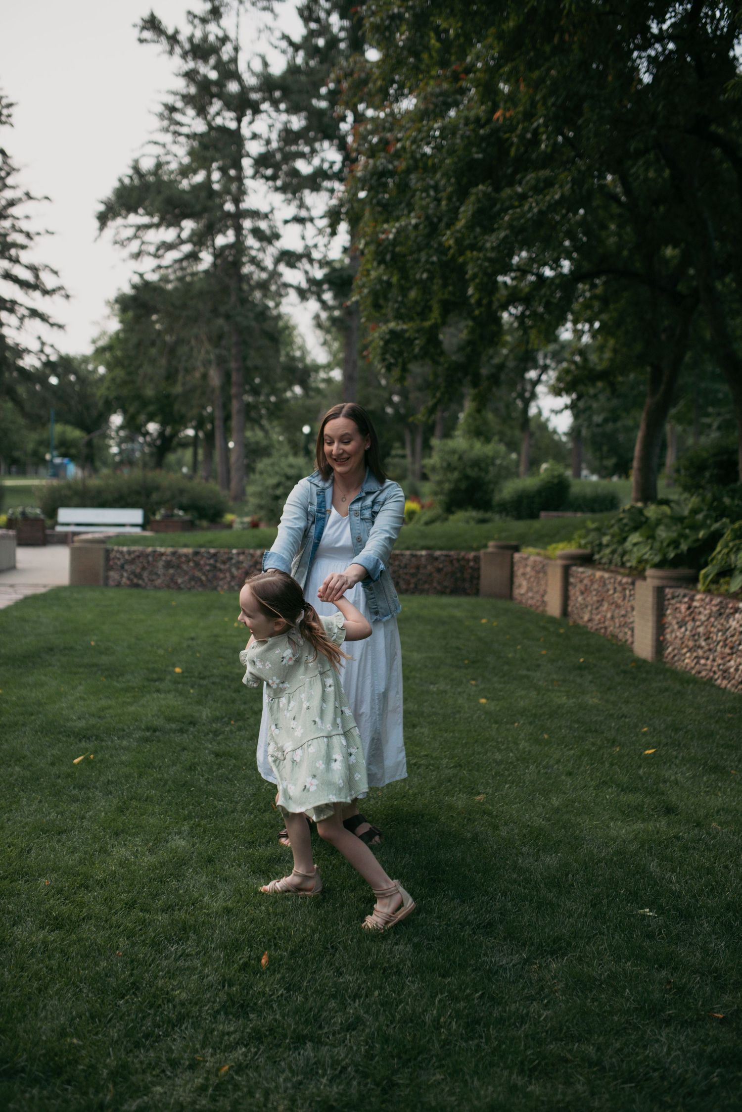 Mother in denim jacket plays with young daughter on green park lawn.