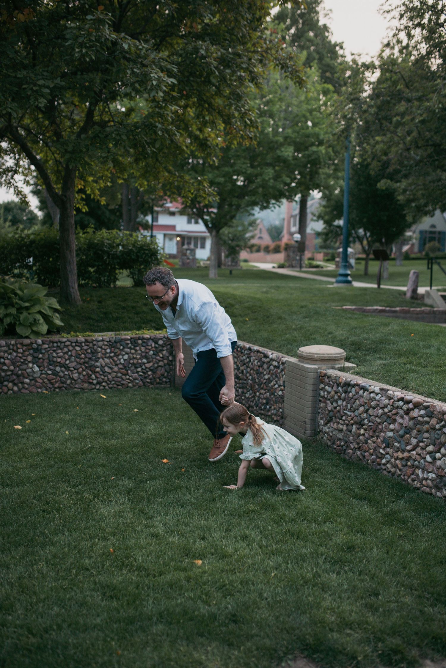 A dad chasing his toddler daughter across a lush green park lawn near a stone wall.