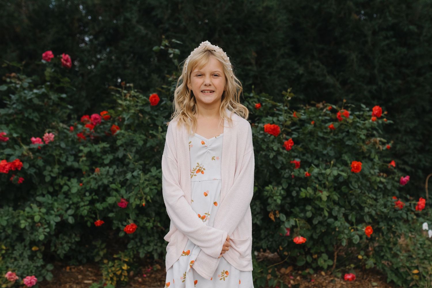 Young blonde girl in a floral dress and pink cardigan standing in front of red rose bushes.