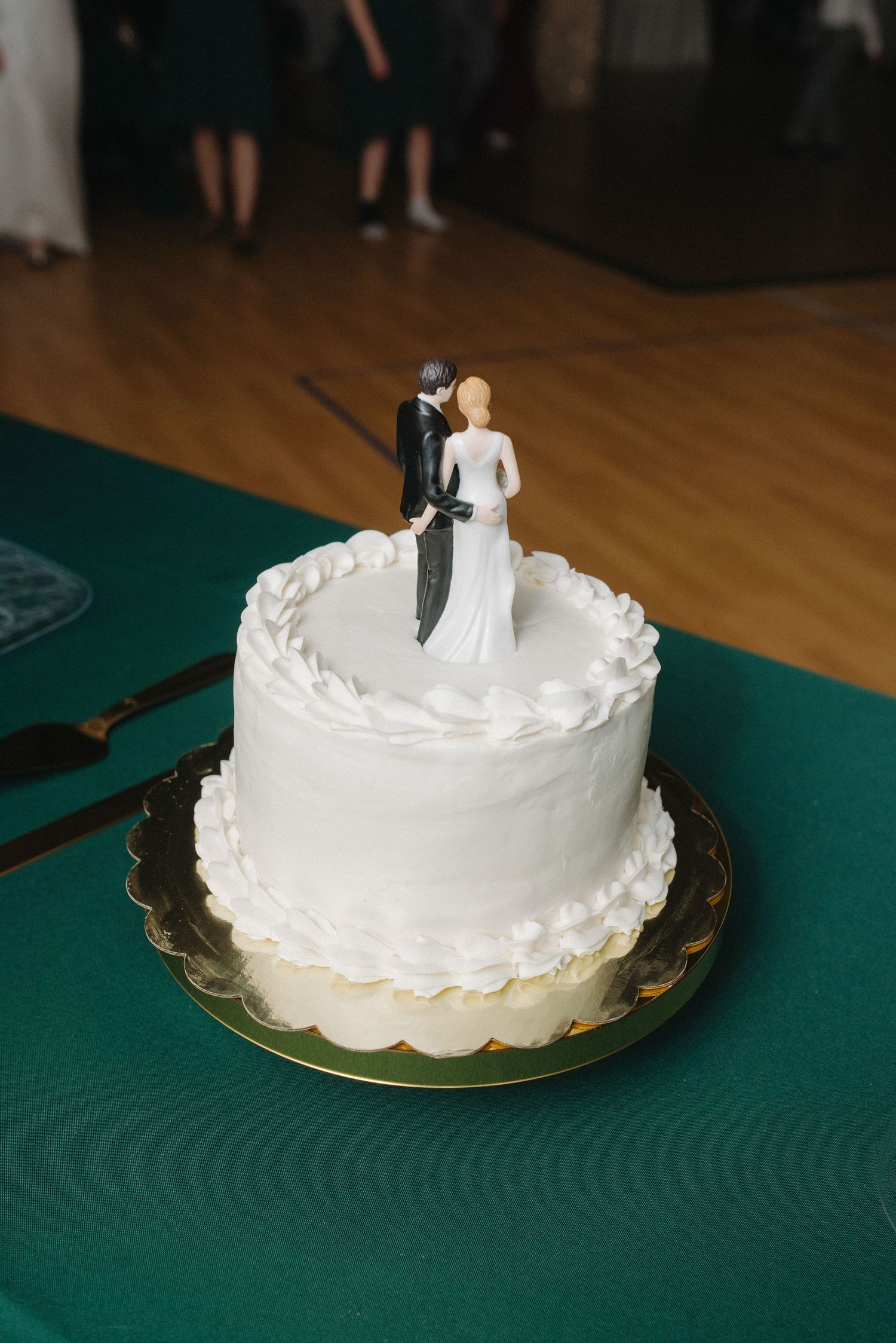 Simple white single-tier wedding cake with classic bride and groom figurine topper on green tablecloth.
