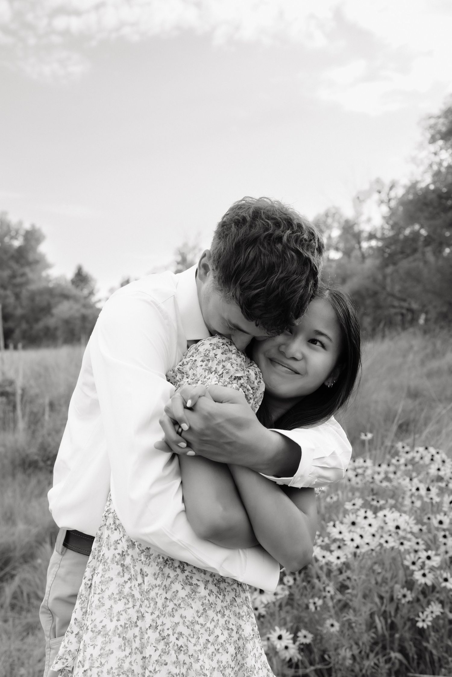 Black and white photo of a young couple embracing in a field, man kissing womans head while she smiles.