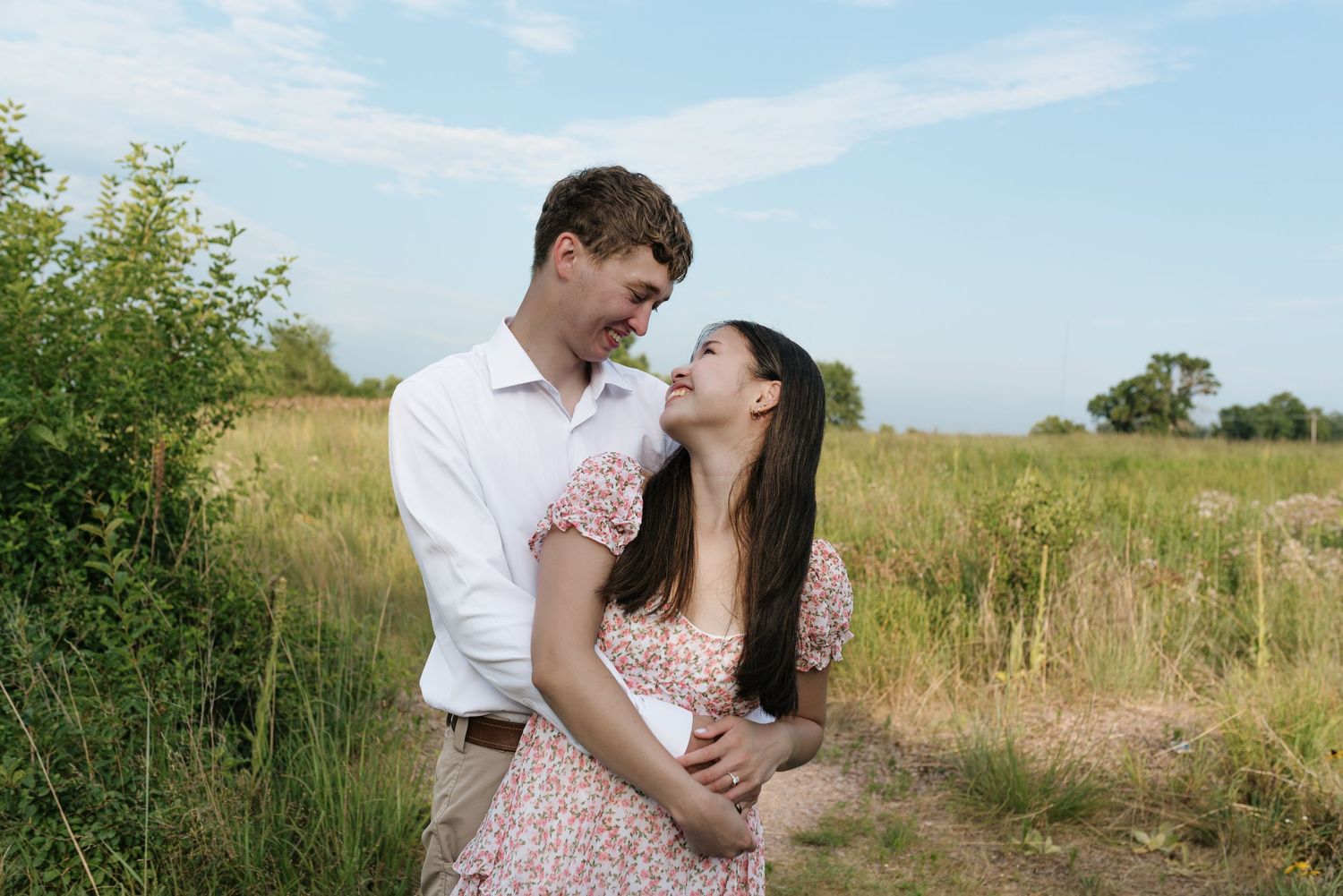 A couple smiles and gazes at each other while embracing on a sunny path in a green meadow.