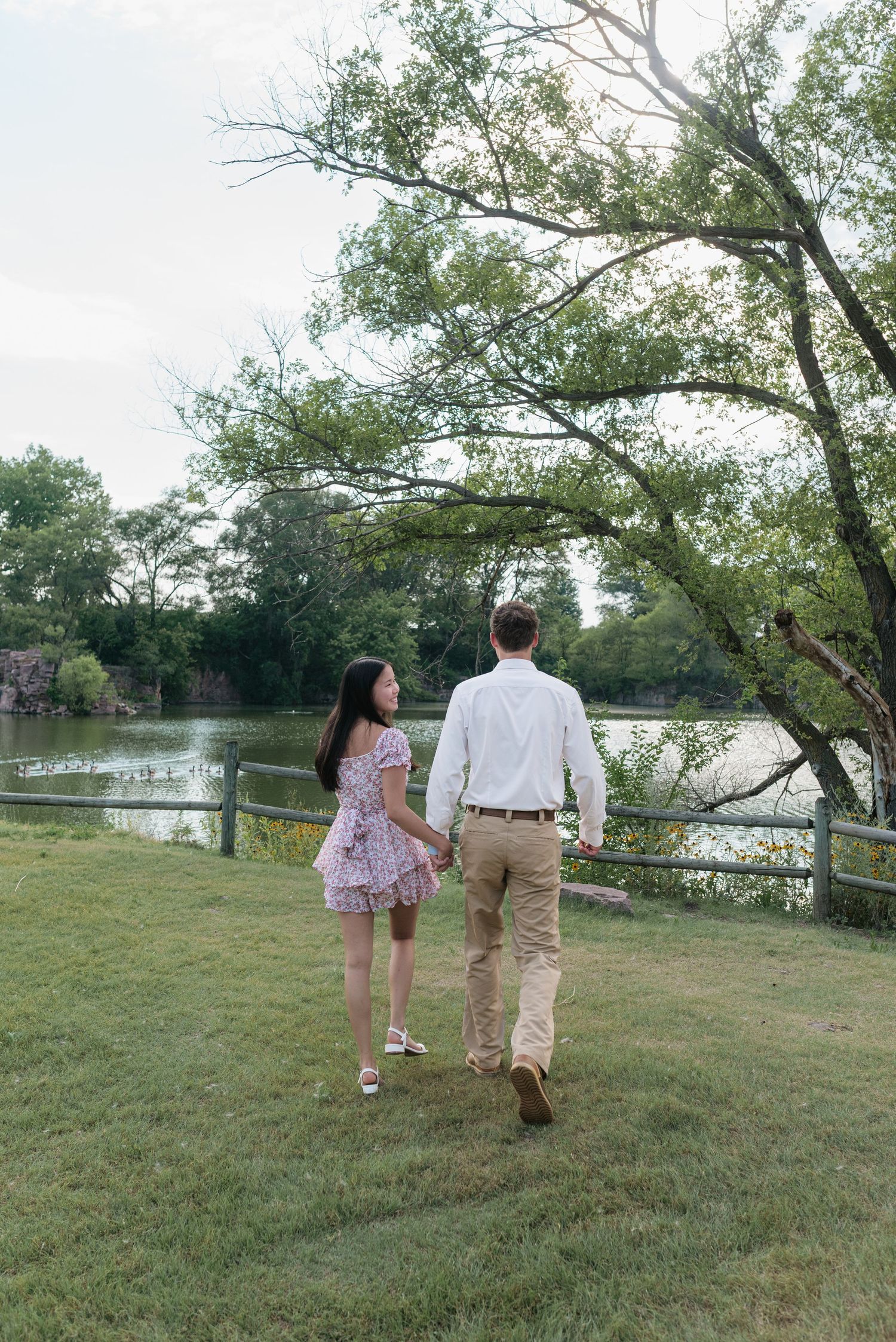 Couple holding hands walking toward a serene pond surrounded by lush green trees and a wooden fence.