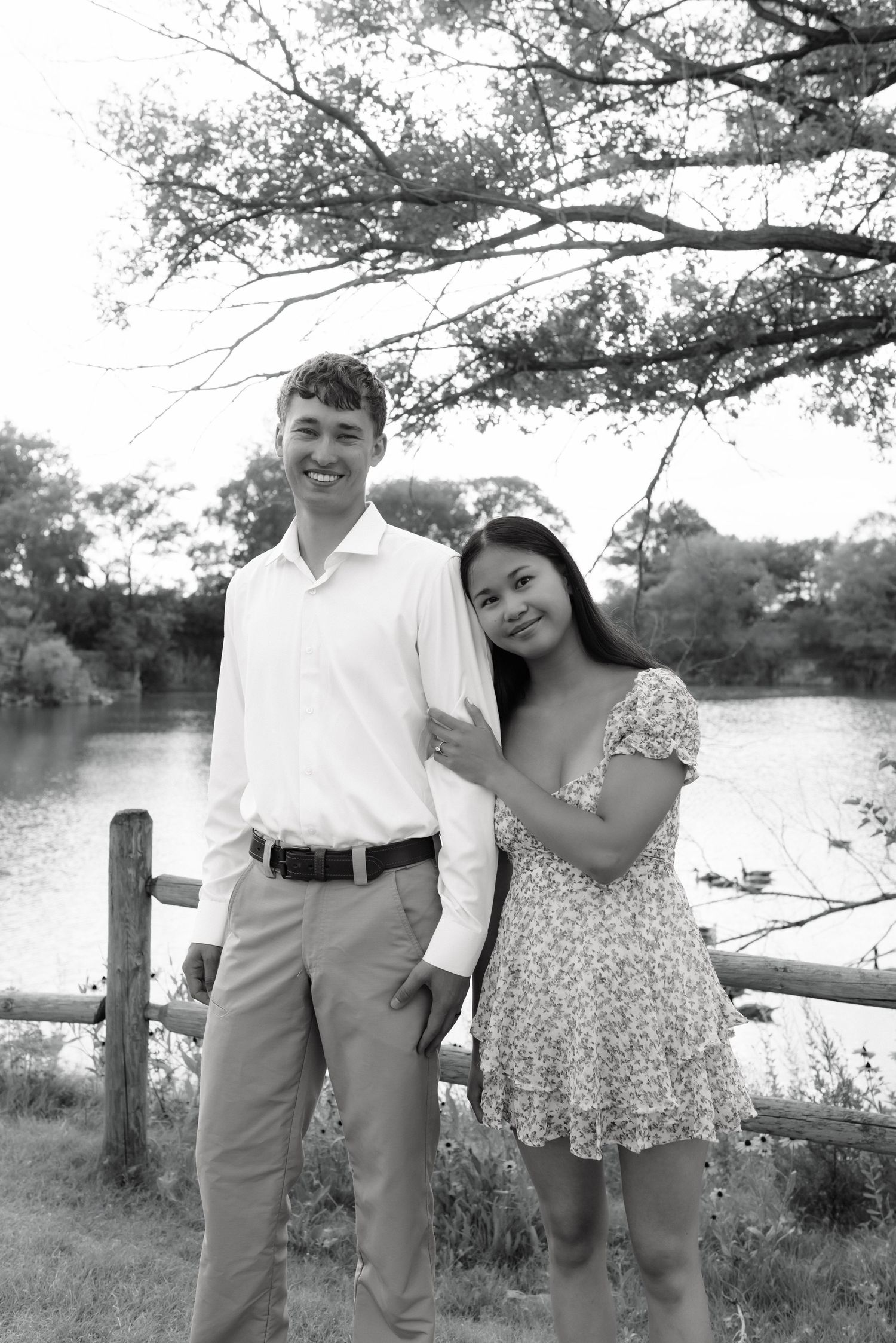A smiling couple poses together in black and white near a rustic fence with a lake backdrop.