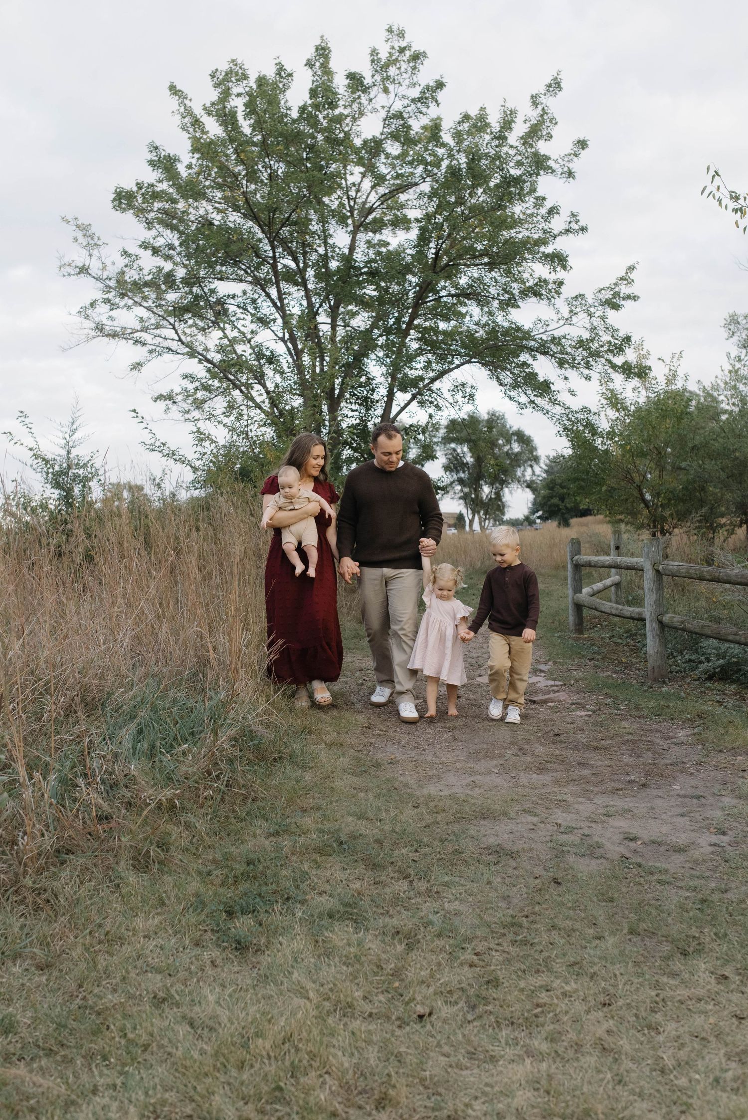 Family walking on rustic dirt path with tall grass, trees, and wooden fence in background.