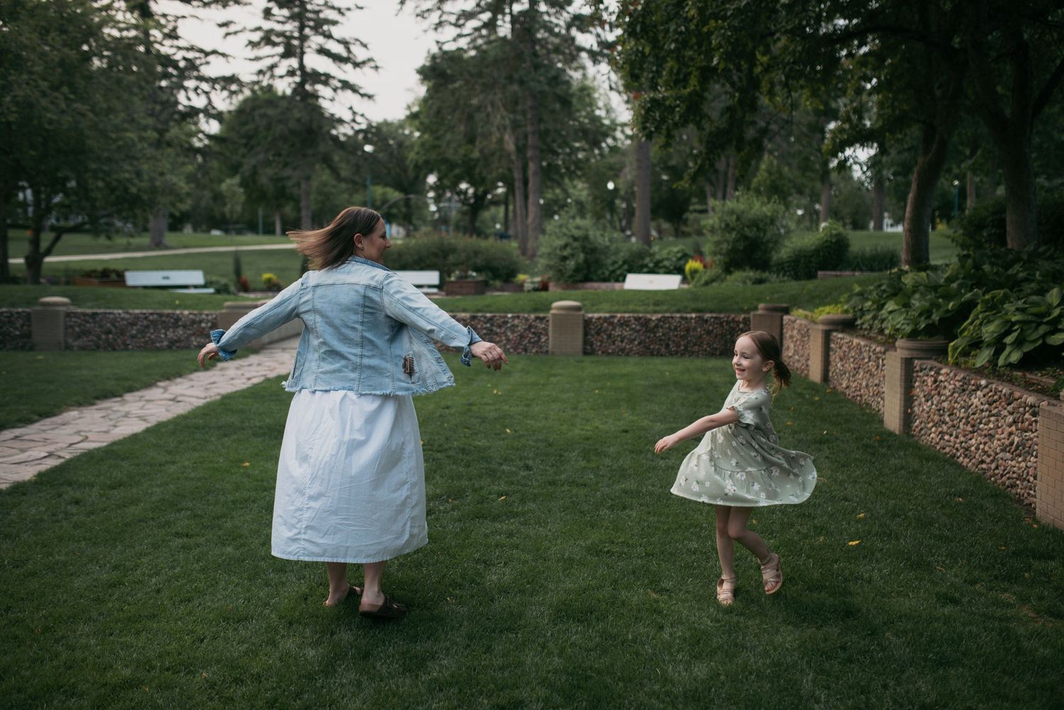 A mother in a white dress and denim jacket spins with her young daughter in a floral dress in a park.