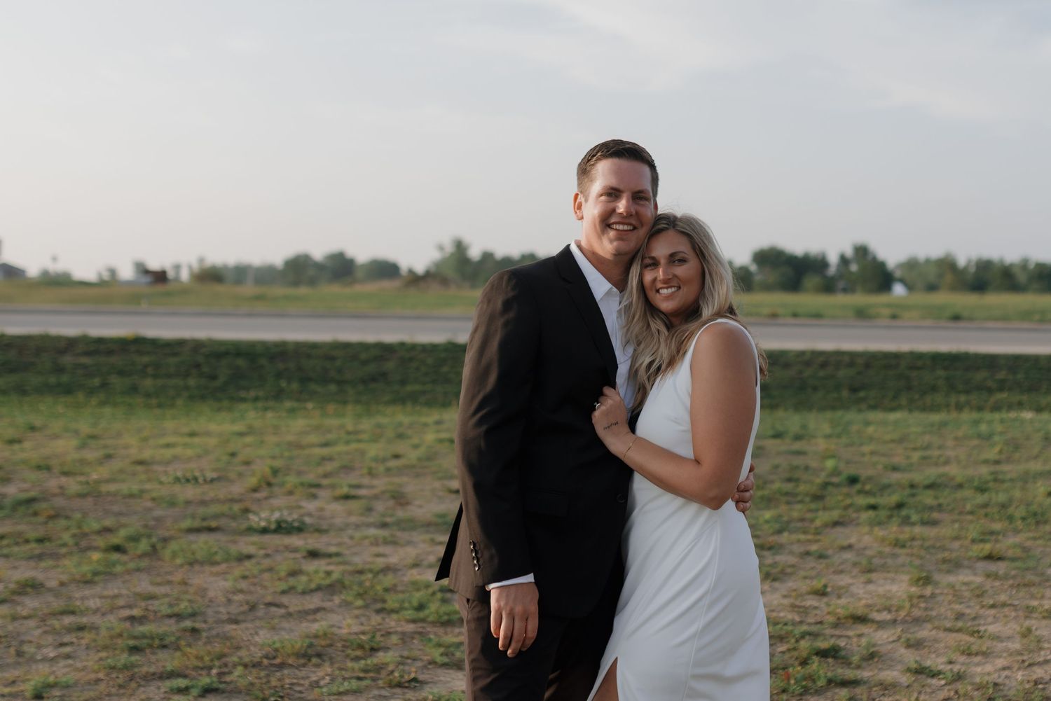 Smiling couple poses together in open field at dusk, bride in white dress and groom in black suit.