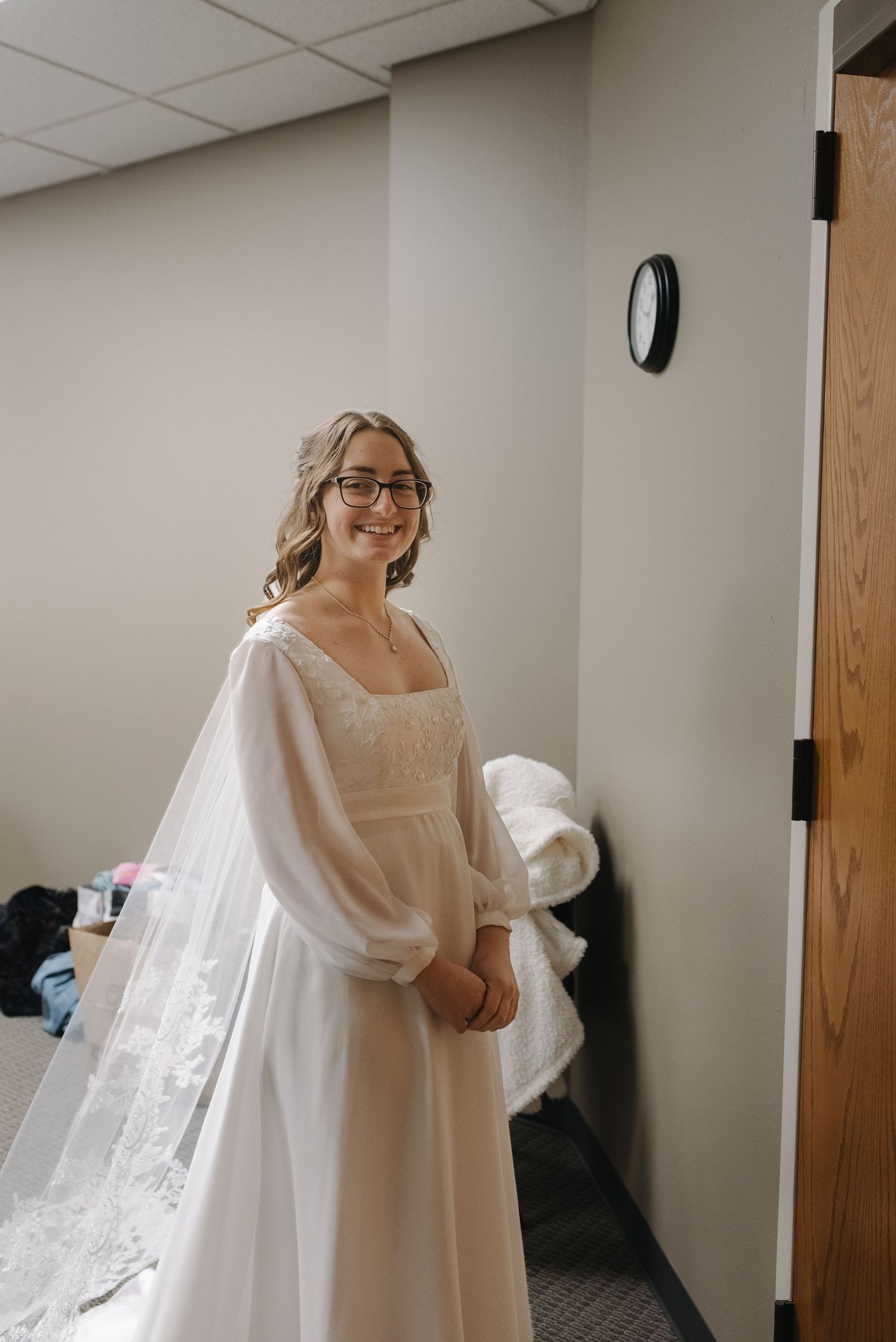 Smiling bride in white lace wedding gown with sheer sleeves and veil, wearing glasses, posing in a bridal suite.