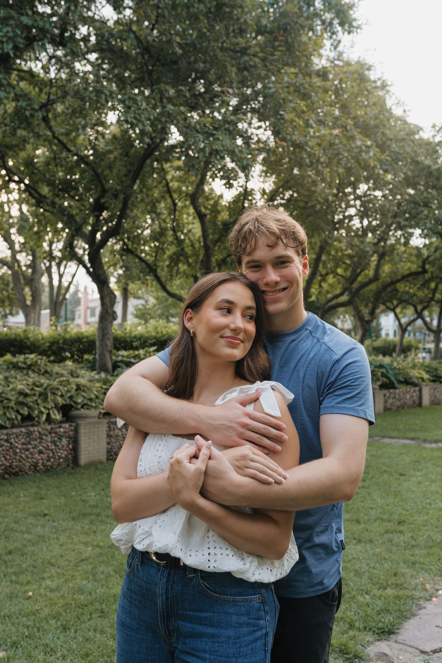 A young couple embracing in a lush green park, smiling warmly, woman in white top and man in blue t-shirt.