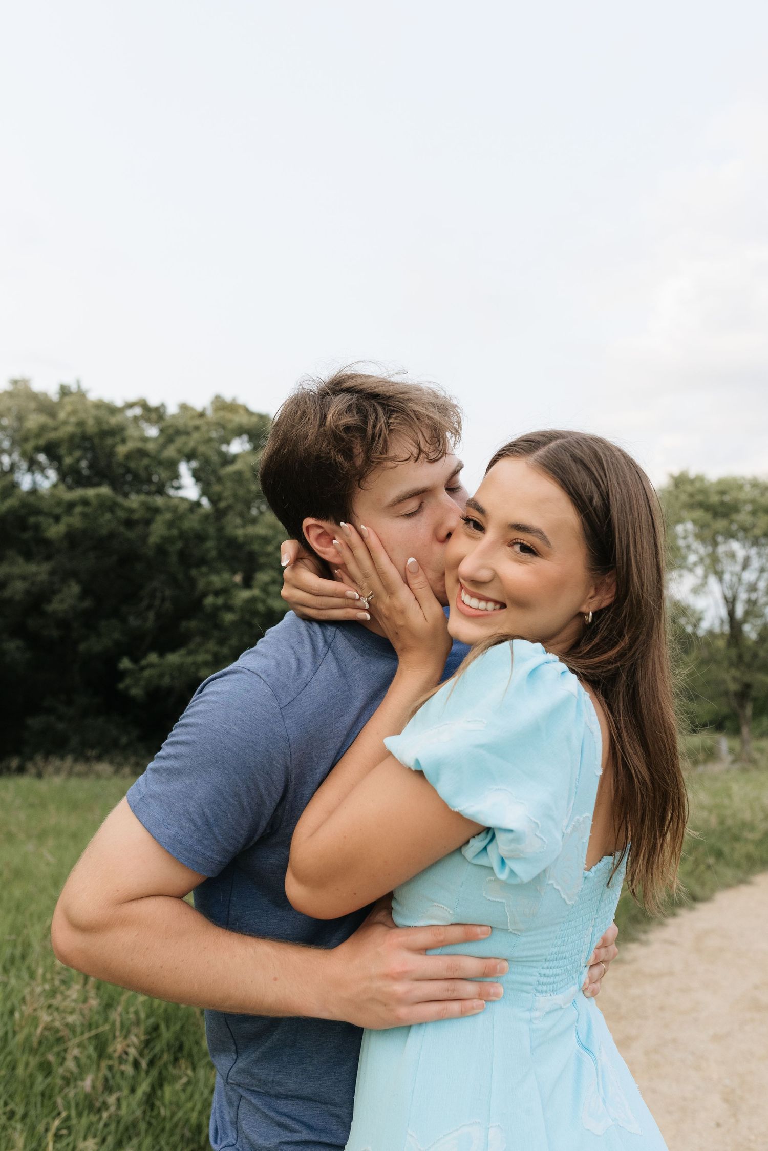 A young couple sharing a romantic kiss outdoors, woman in light blue dress smiling, man in blue t-shirt embracing her.