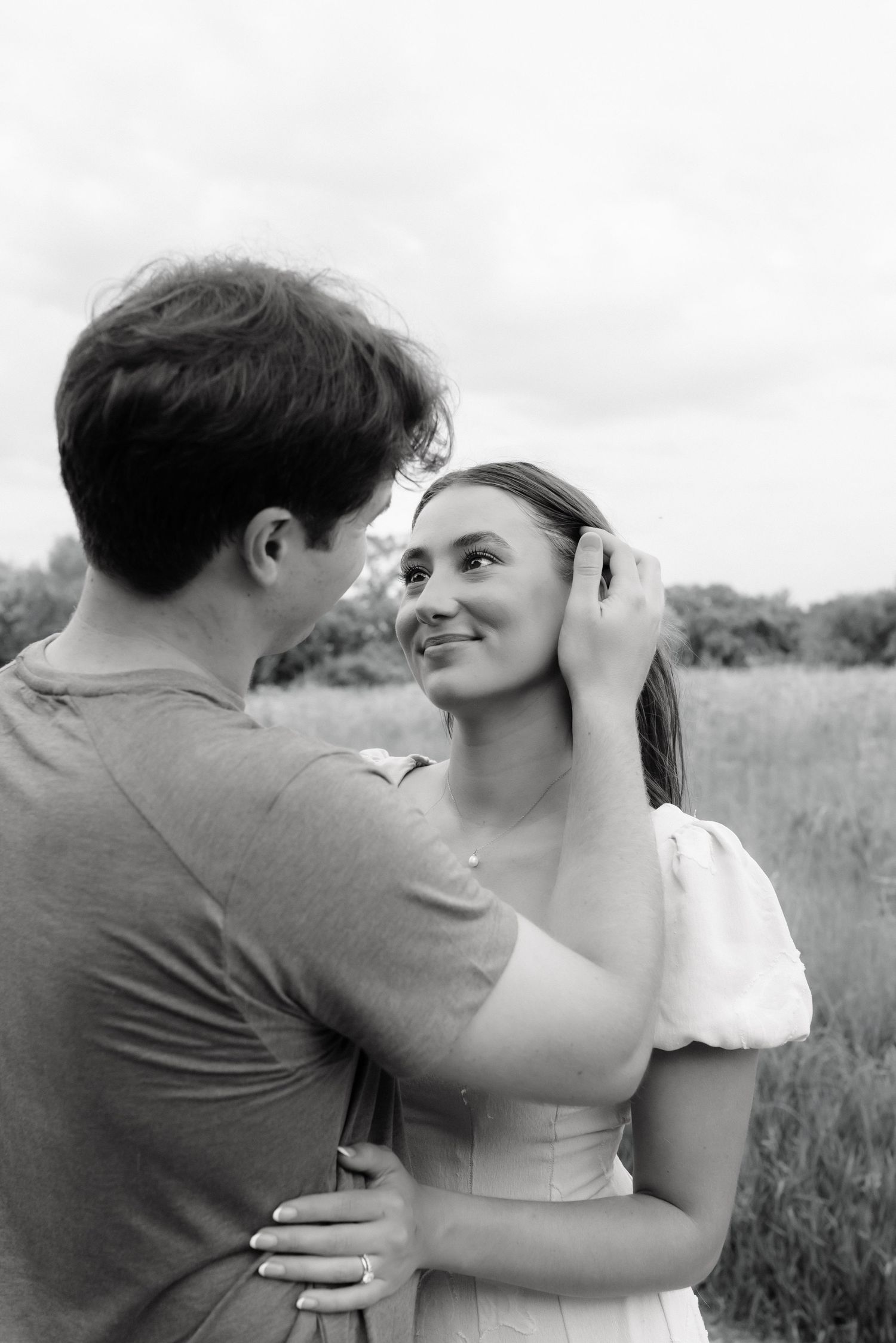 Black and white photo of a couple embracing in a field, man tucking womans hair back as she smiles up at him.