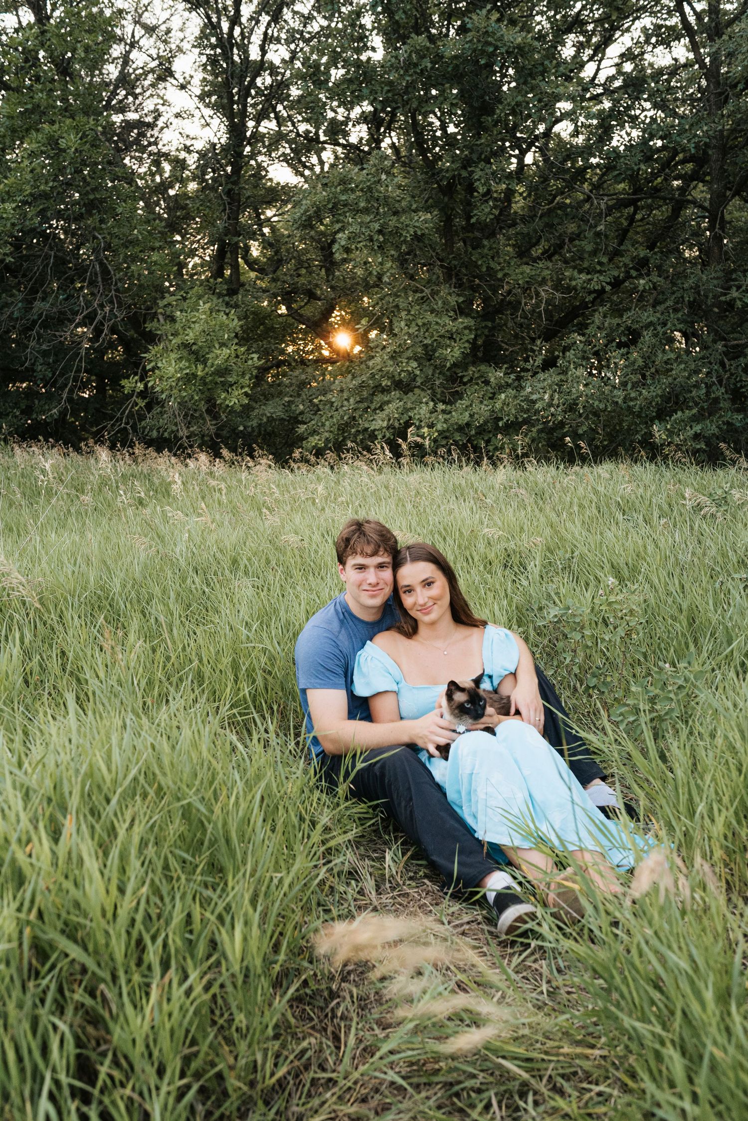 Couple sitting in lush green field at sunset, holding a small dog, wearing casual blue outfits with trees in background.