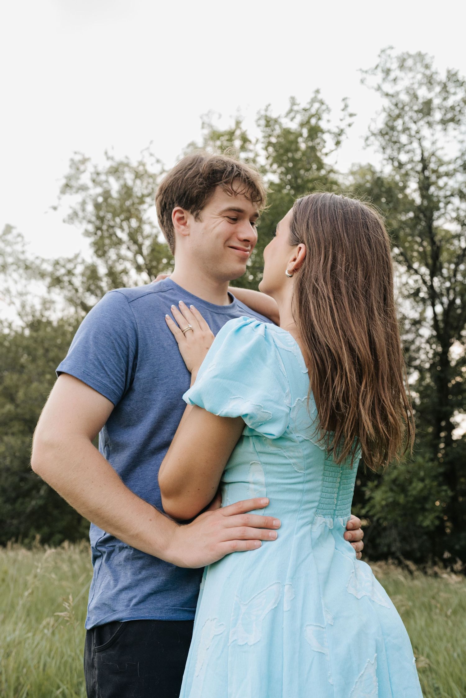 A young couple embraces in a green field, the woman in a light blue dress gazing at a smiling man in a blue t-shirt.