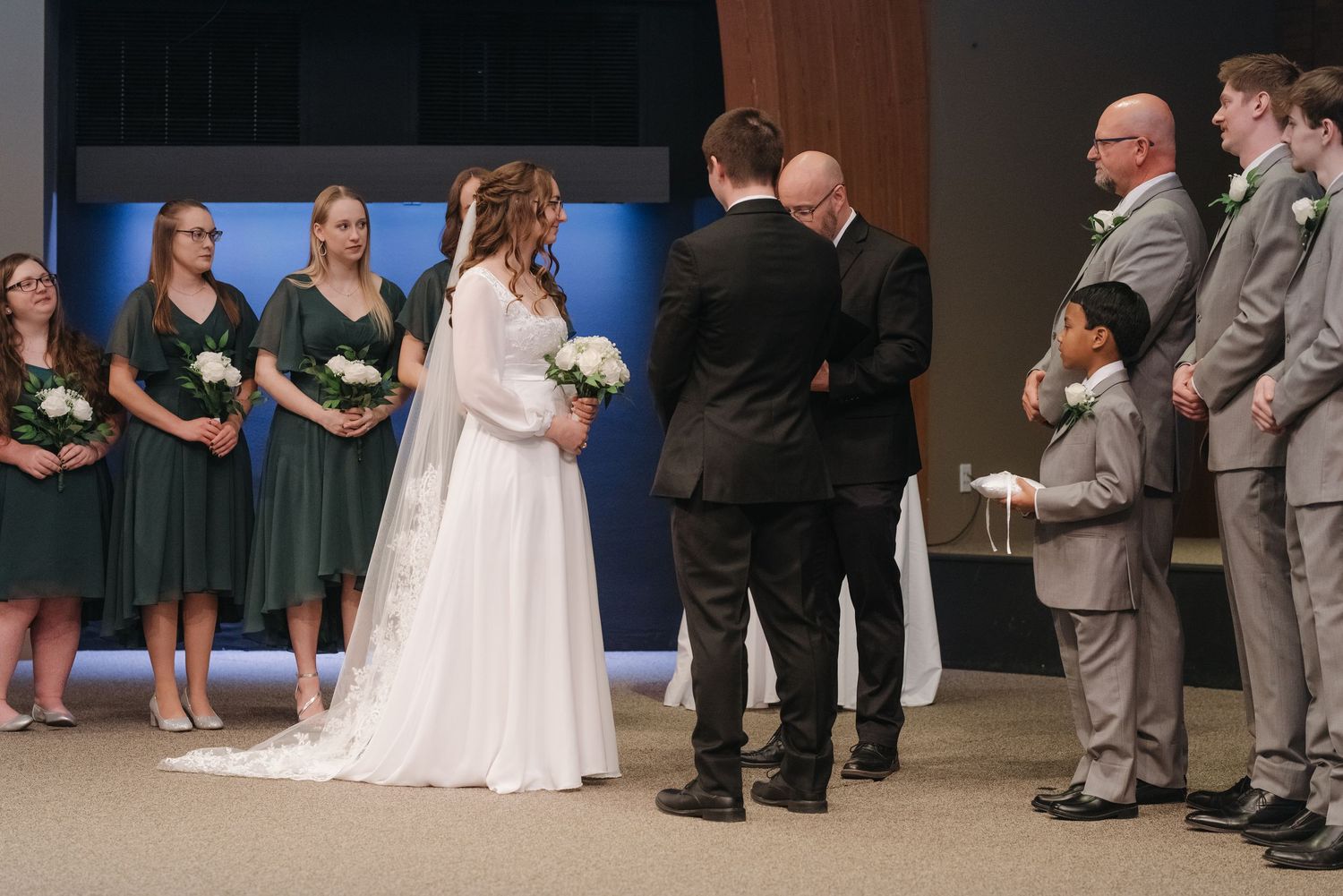 Wedding ceremony with bride in white gown, groom, officiant, bridesmaids in green dresses, and groomsmen in gray suits.