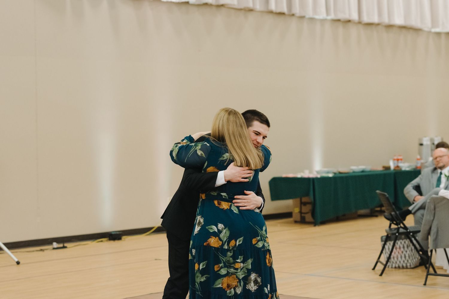 Groom in black suit sharing warm embrace with woman in floral dress on wedding dance floor.