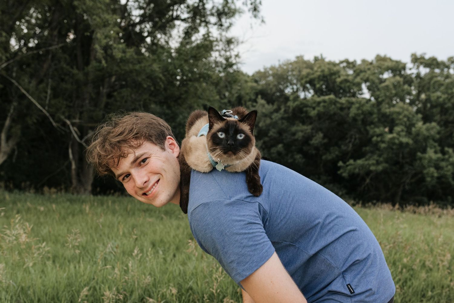 Young man smiling outdoors with a Siamese cat perched on his shoulders in a green grassy field.
