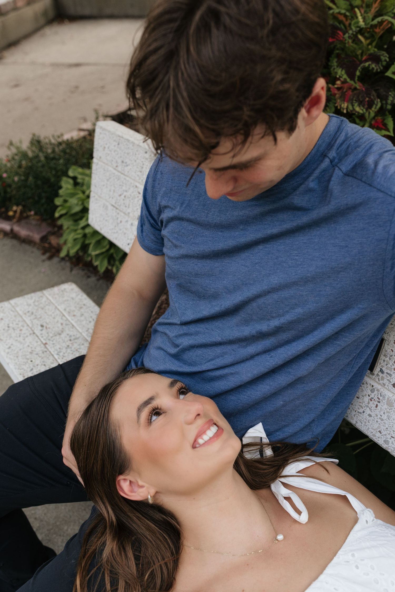 A young woman lying on a bench gazes up smiling at a man in a blue t-shirt looking down at her outdoors.