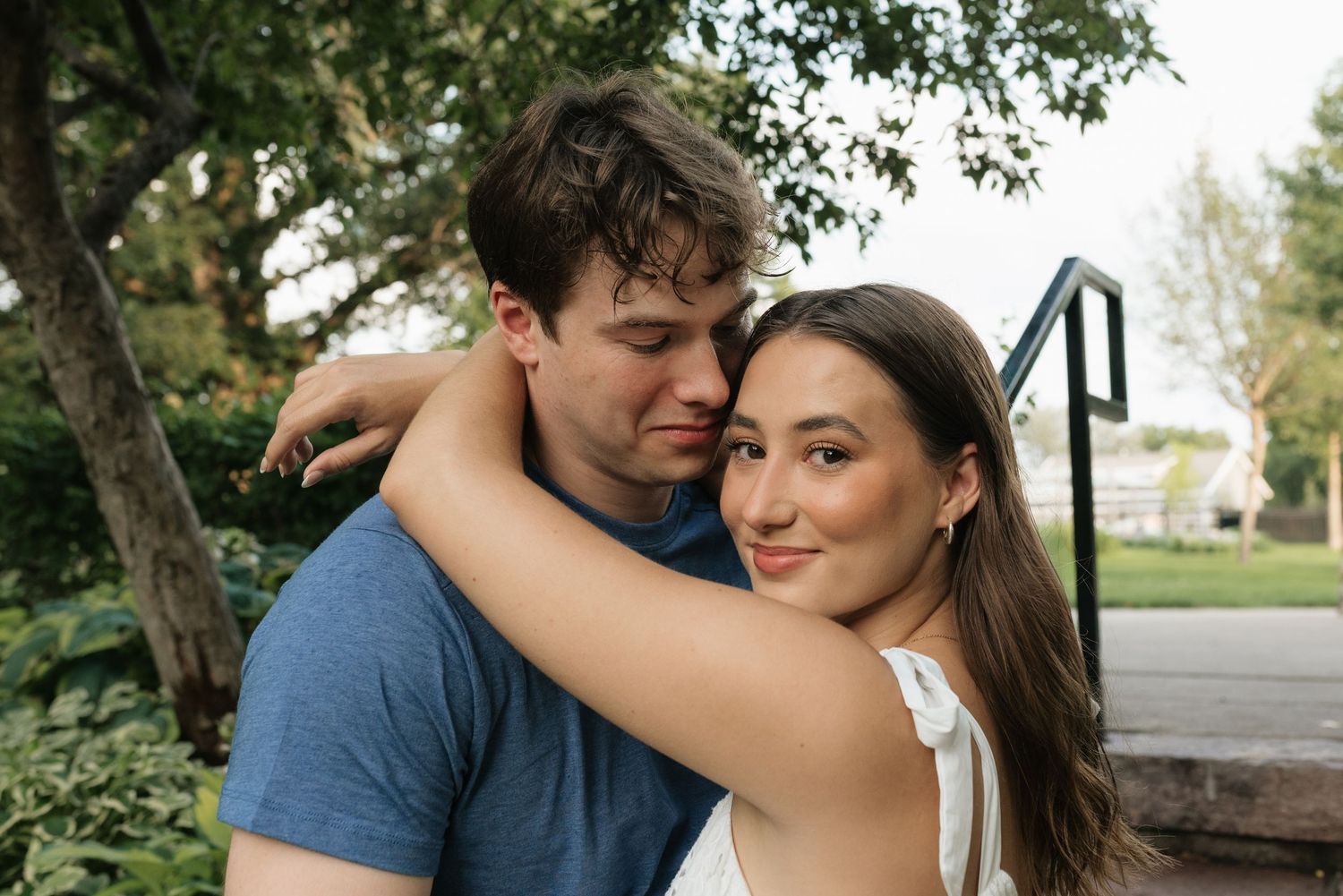 A young couple embracing outdoors, woman smiling at camera while hugging man near green trees and steps.