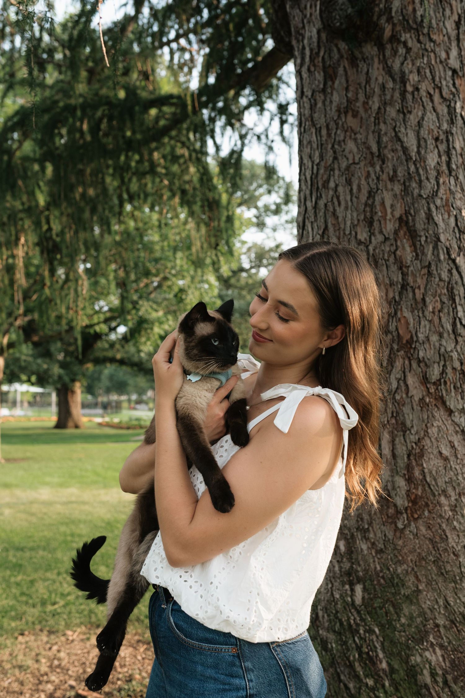Young woman in white top holding a Siamese cat outdoors near a large tree in a lush green park.