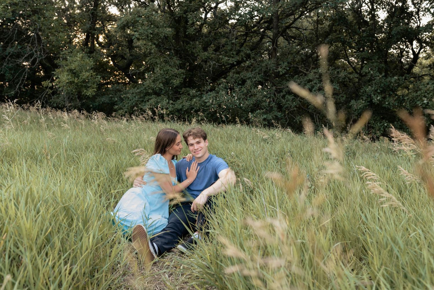 Couple sitting together in tall green grass field surrounded by trees during golden hour outdoor photo session.