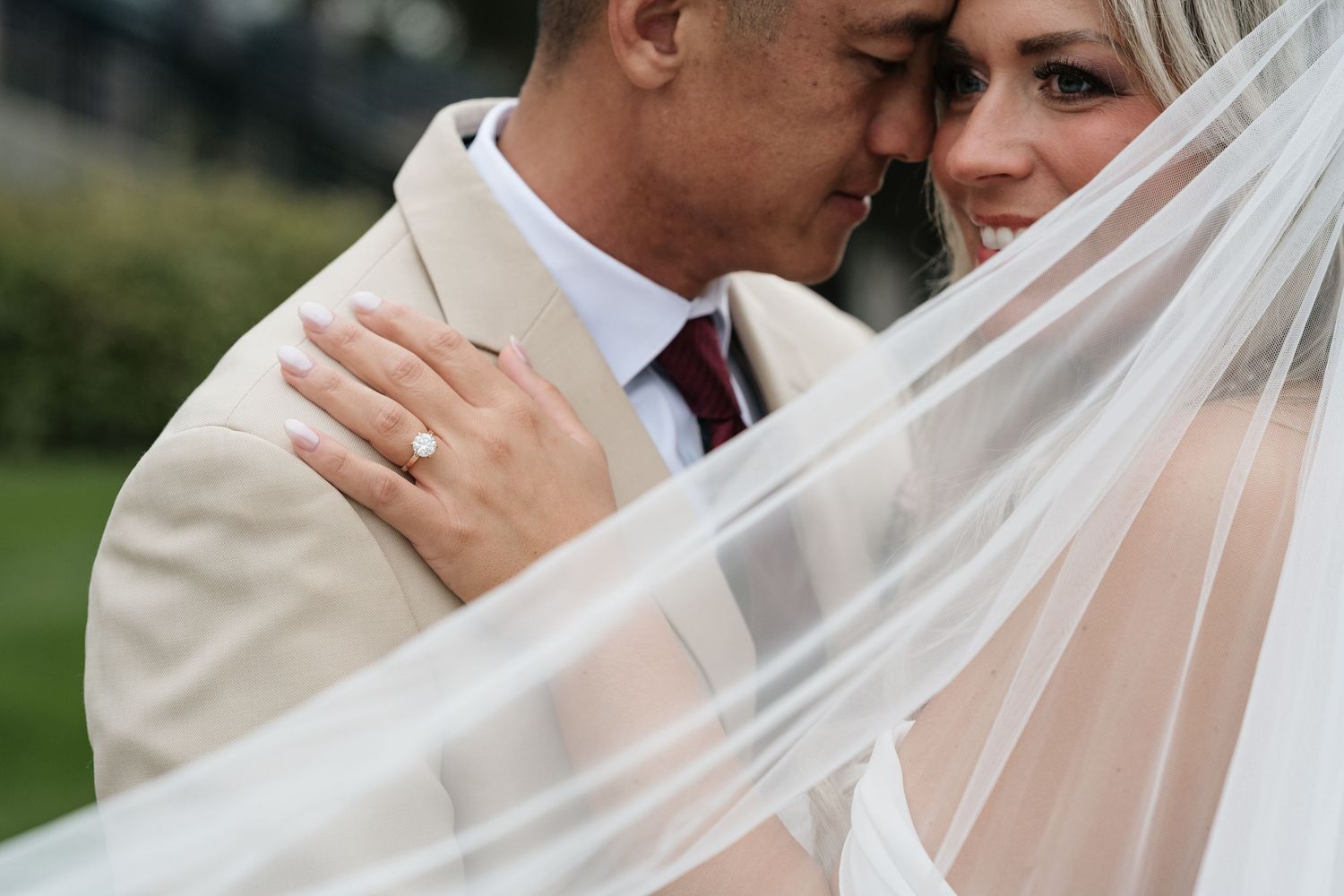 Bride and groom share an intimate moment under a flowing white veil, with the groom in a beige suit and burgundy tie.