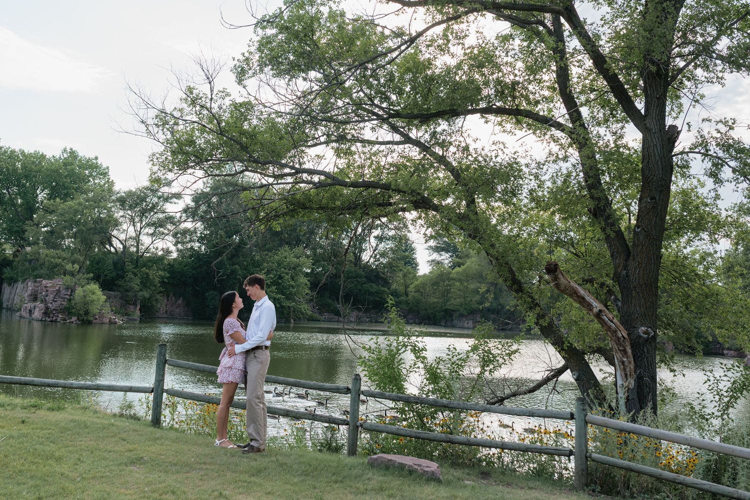 Couple embracing near a wooden fence by a serene lake surrounded by lush green trees in a romantic outdoor setting.