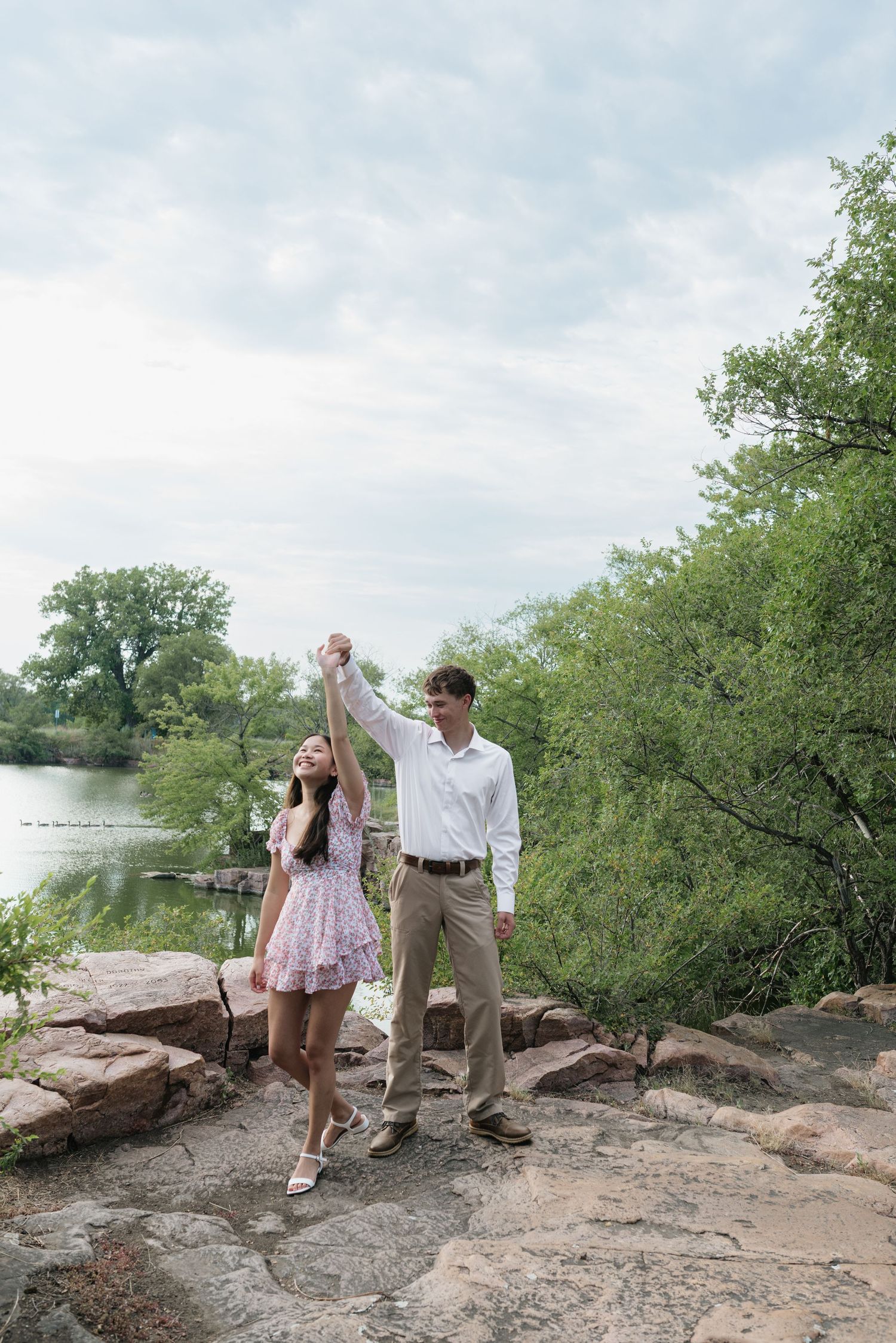 Couple holding hands by a lake, man twirling woman in pink floral dress on rocky shore.