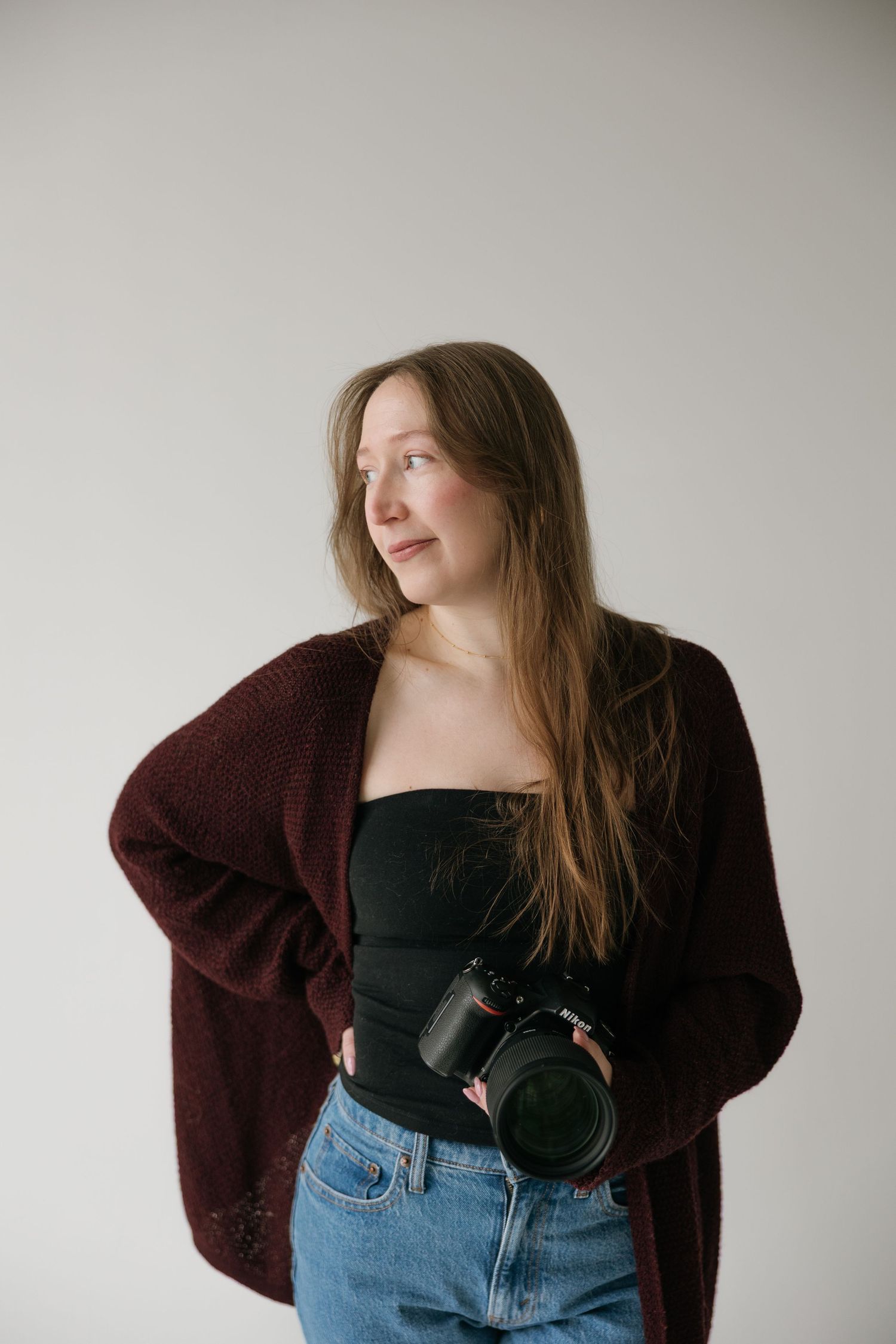 Female photographer holding camera wearing burgundy cardigan and jeans in studio setting.