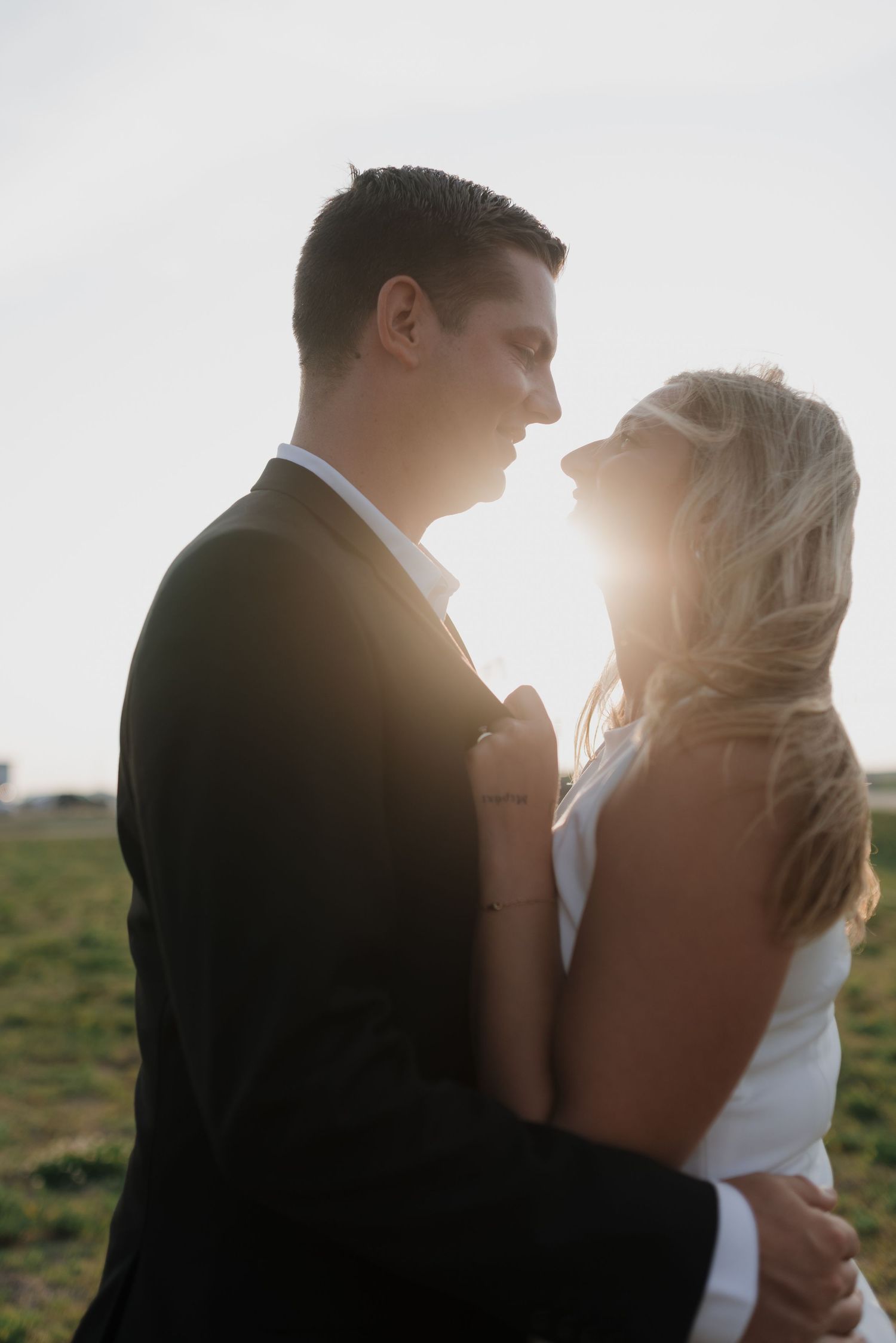 Romantic couple in wedding attire holding each other close with warm sunset light shining between them.