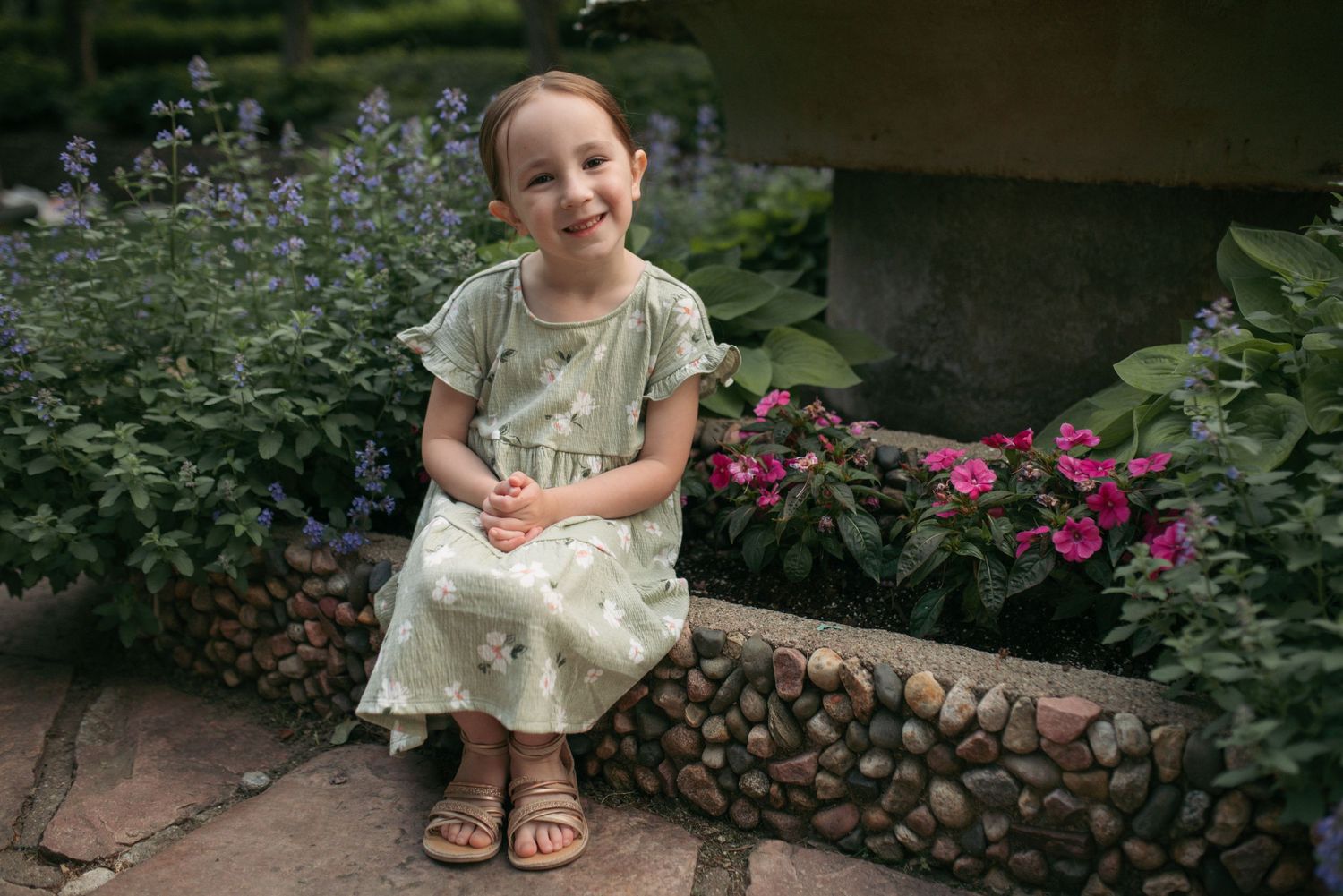 Smiling little girl in green floral dress sits by colorful garden flowers.
