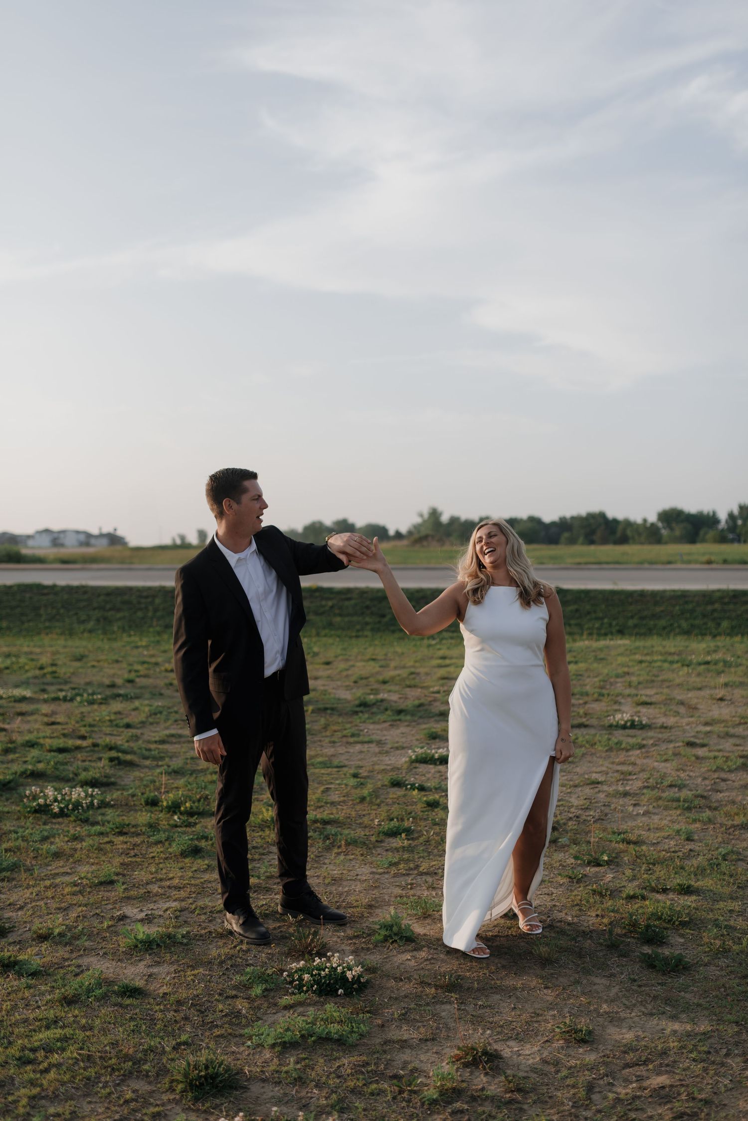 Couple laughing and holding hands outdoors in field, man in suit reaching toward woman in white dress.
