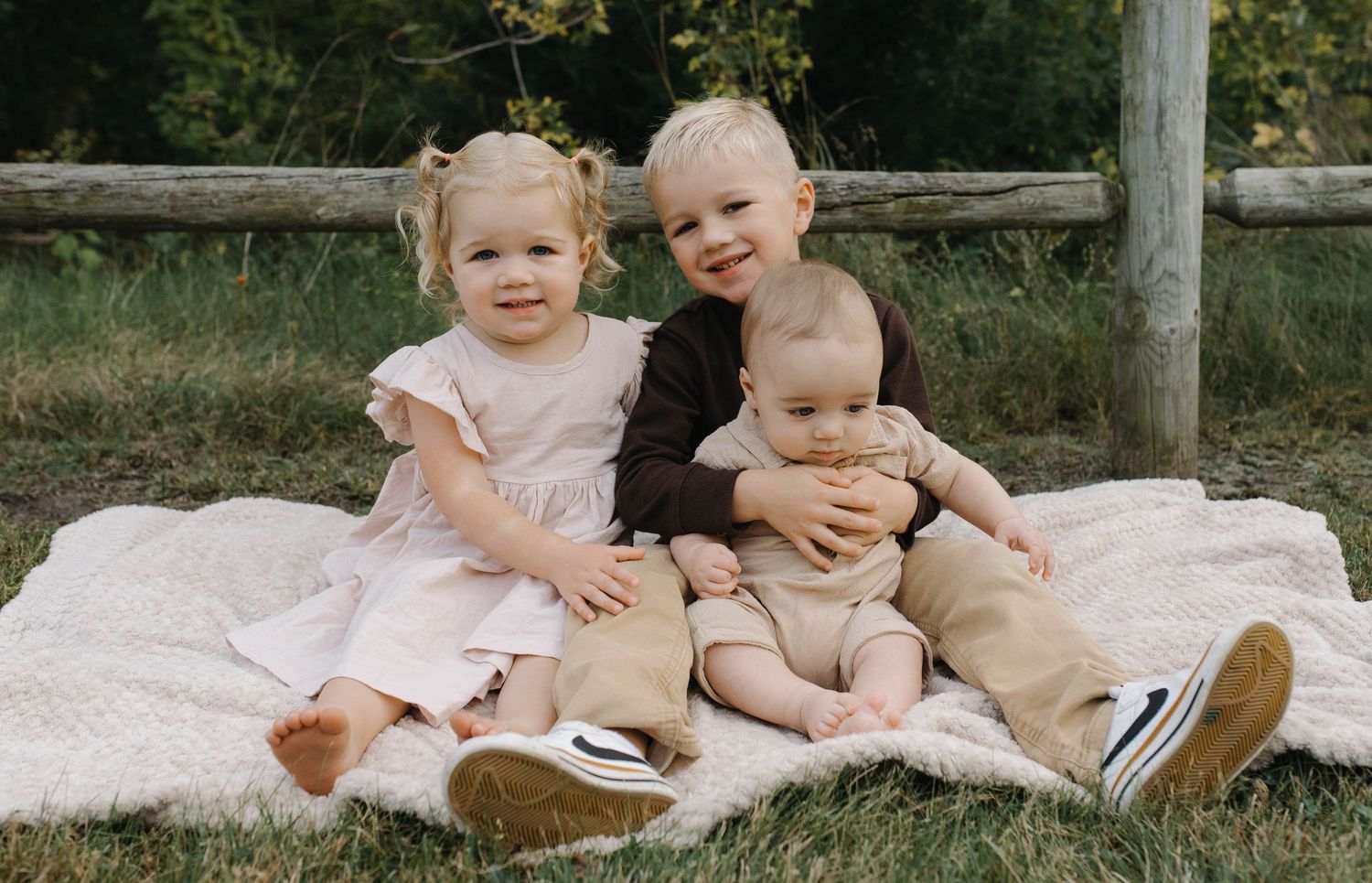 Adorable group of three young siblings sitting on a blanket for an outdoor family photo session in nature.