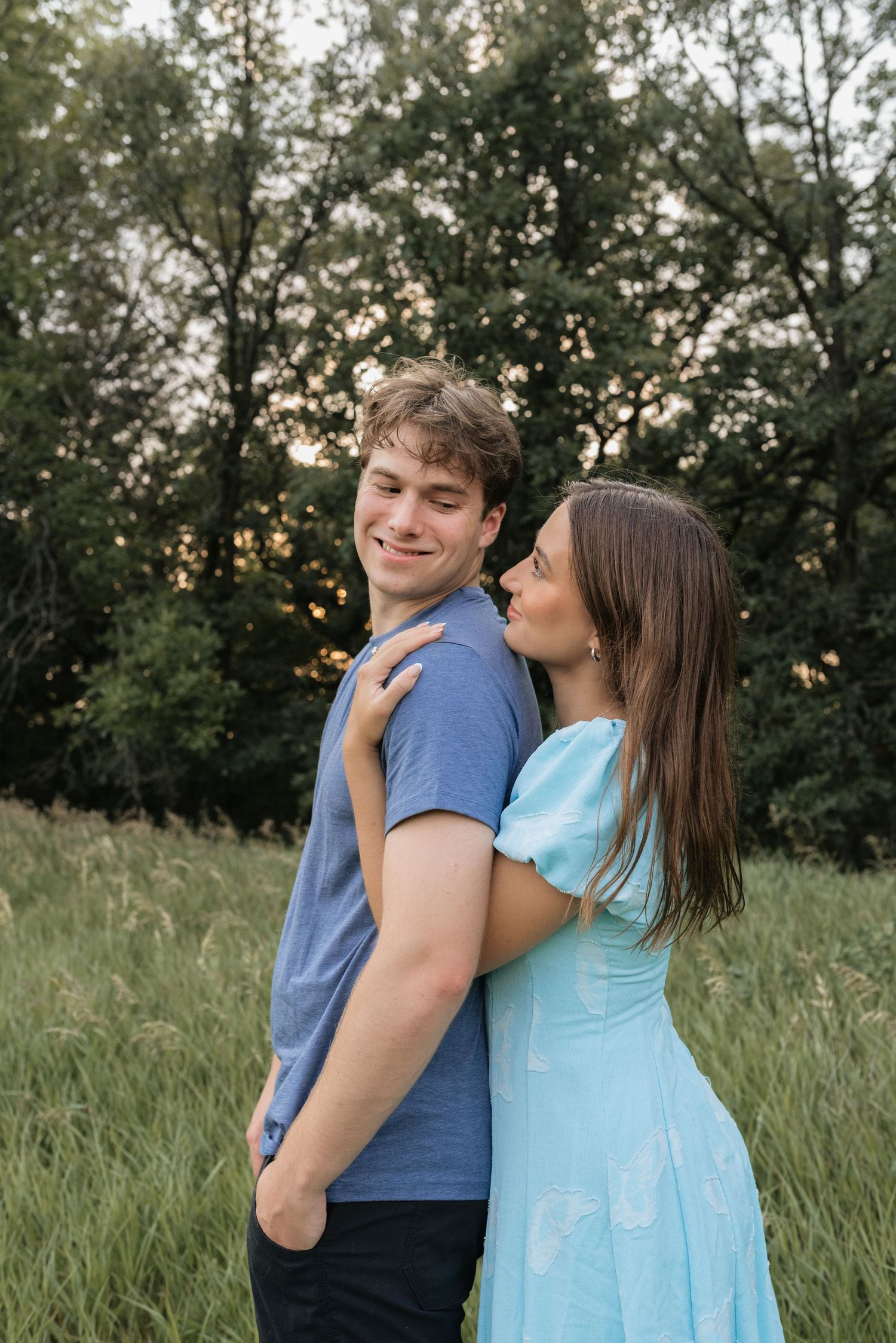 A couple embraces in a green meadow, woman in blue dress kissing man's cheek as he smiles softly.