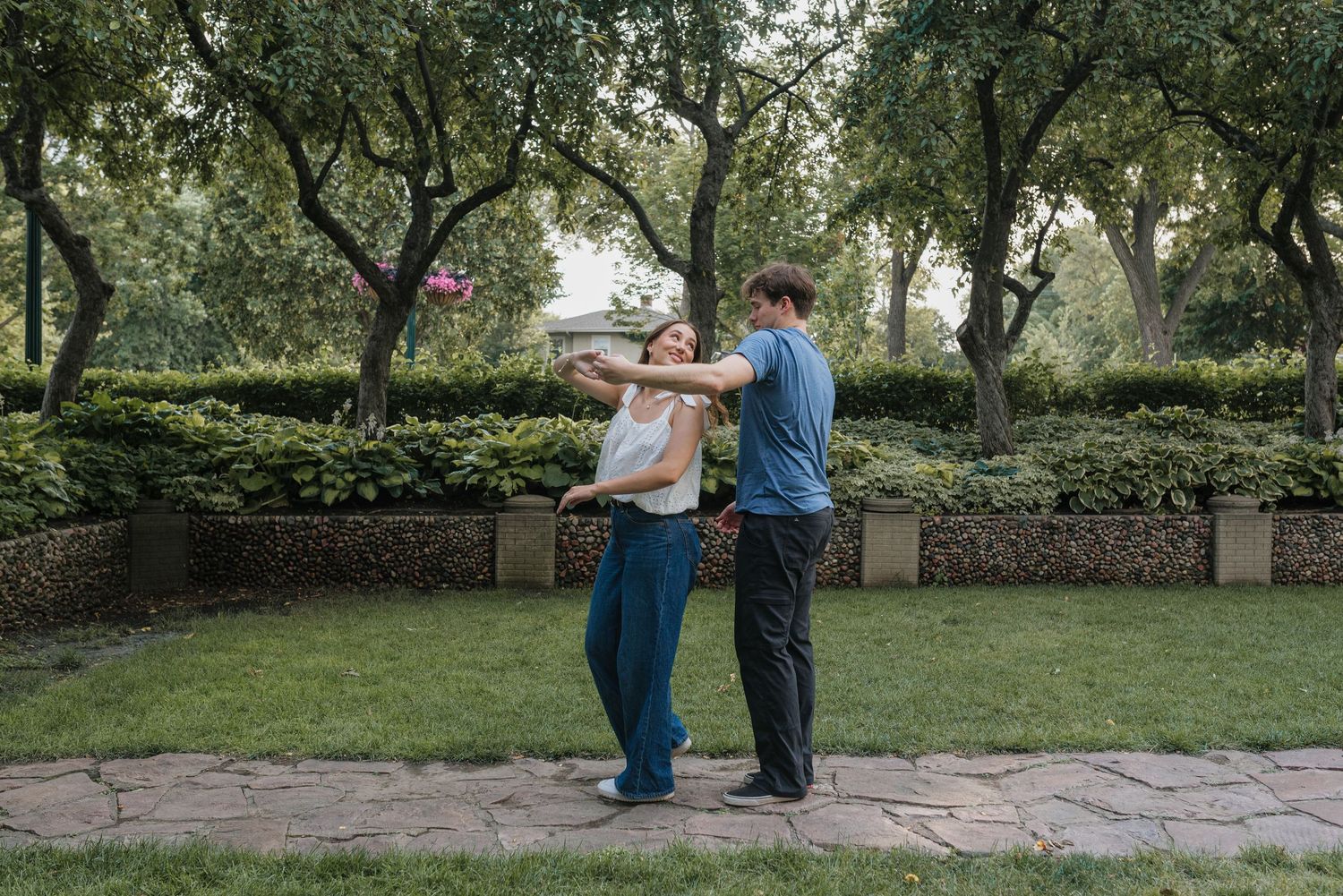 A couple dances together joyfully in a lush green park surrounded by trees and hedges.