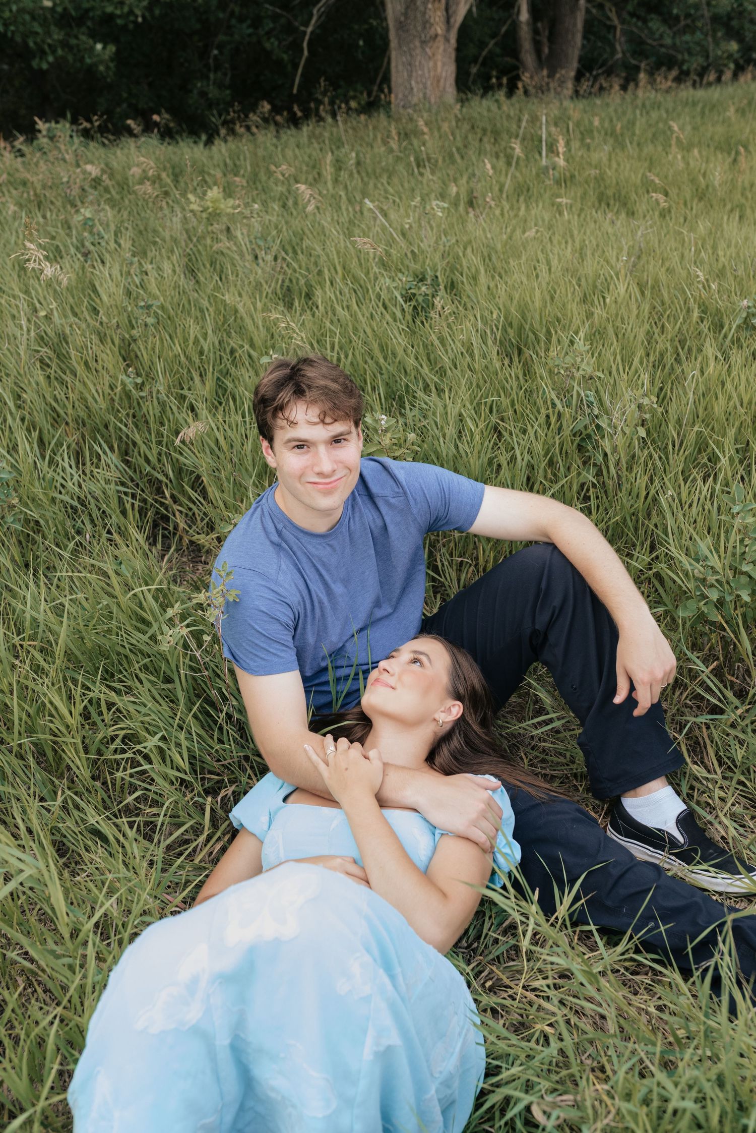 Young couple relaxing in tall green grass field, woman lying in man's lap, both smiling, wearing blue outfits.
