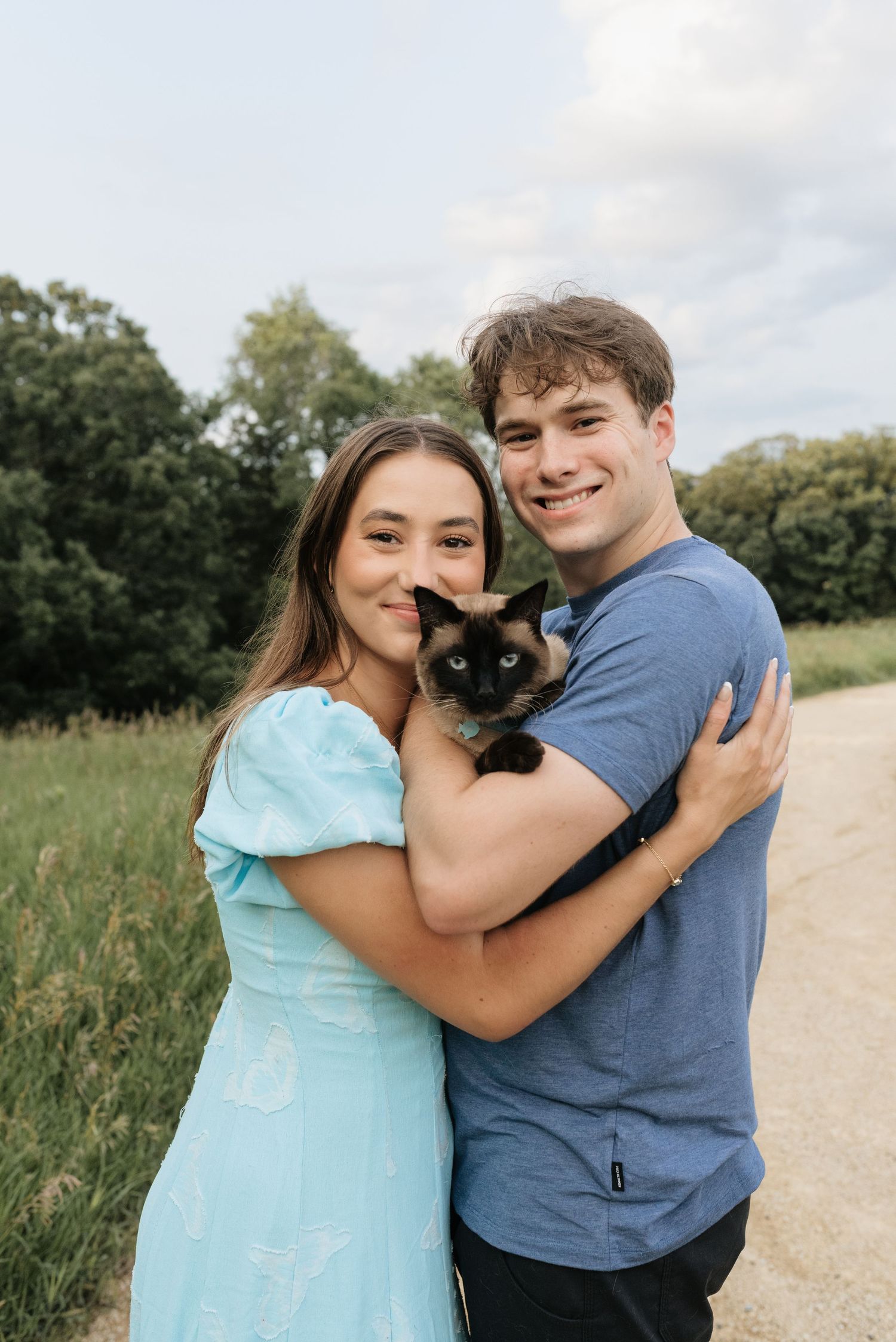 A smiling couple outdoors holding a Siamese cat, woman in light blue dress, man in blue t-shirt, green trees behind.
