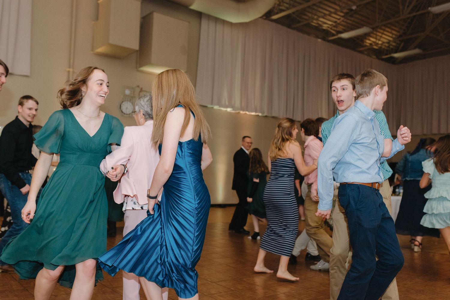Wedding guests in teal and blue dresses dancing joyfully together at a lively wedding reception.