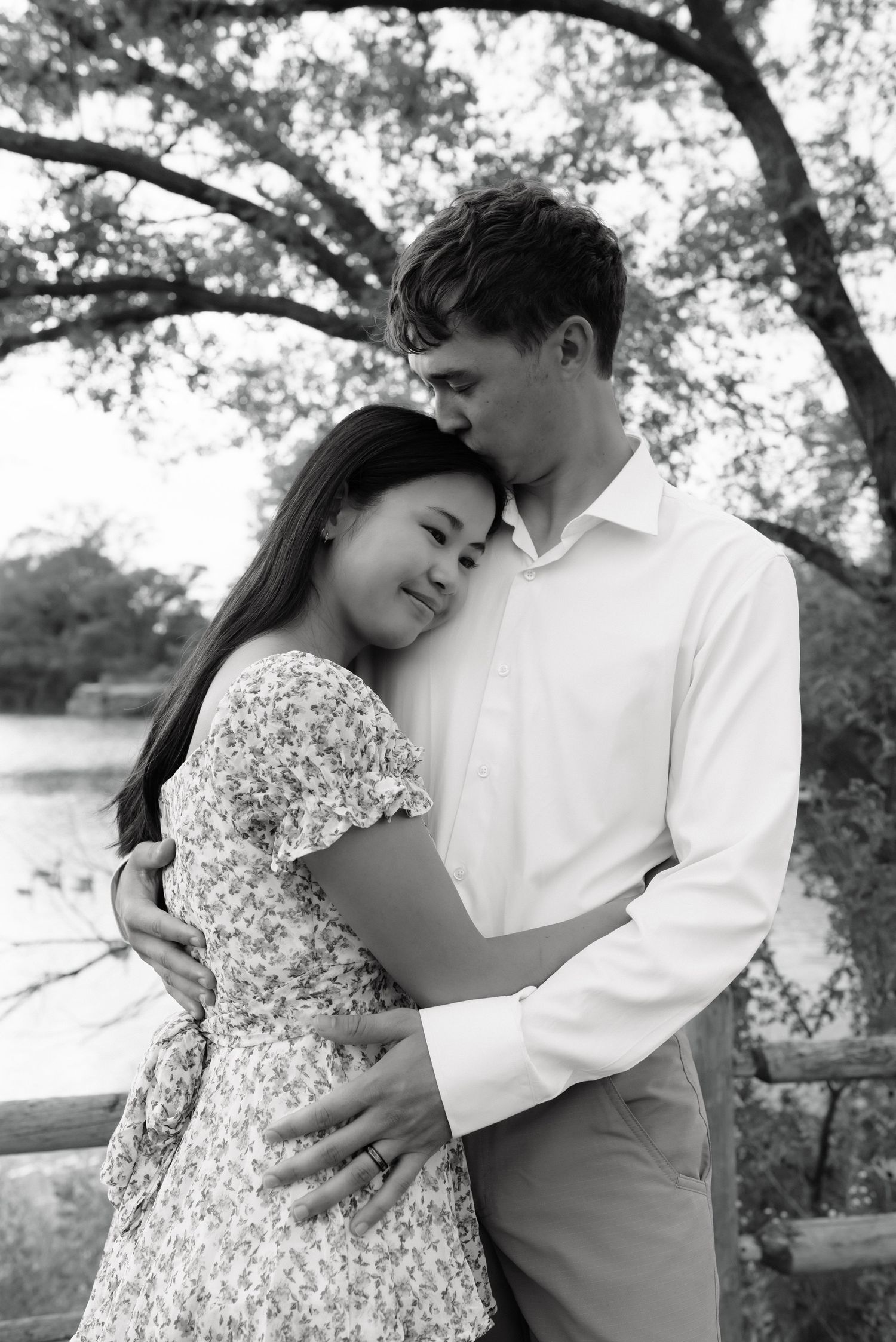 Black and white photo of a couple embracing tenderly near a wooden fence surrounded by trees.