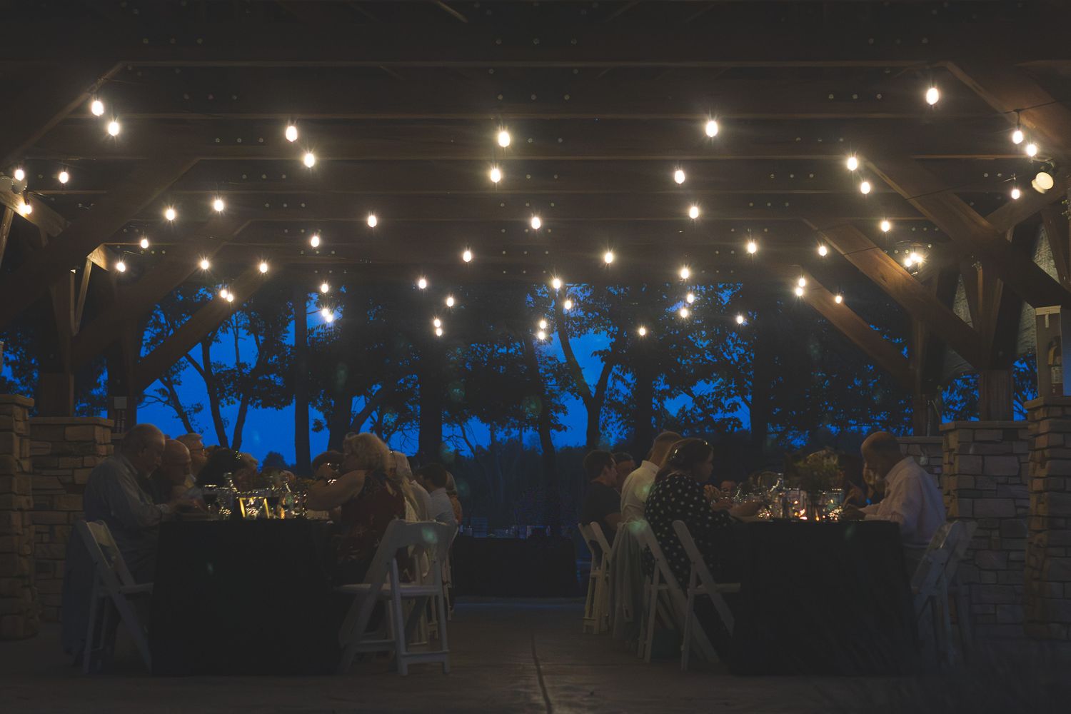 Outdoor evening reception with string lights illuminating guests dining under a rustic wooden pergola.