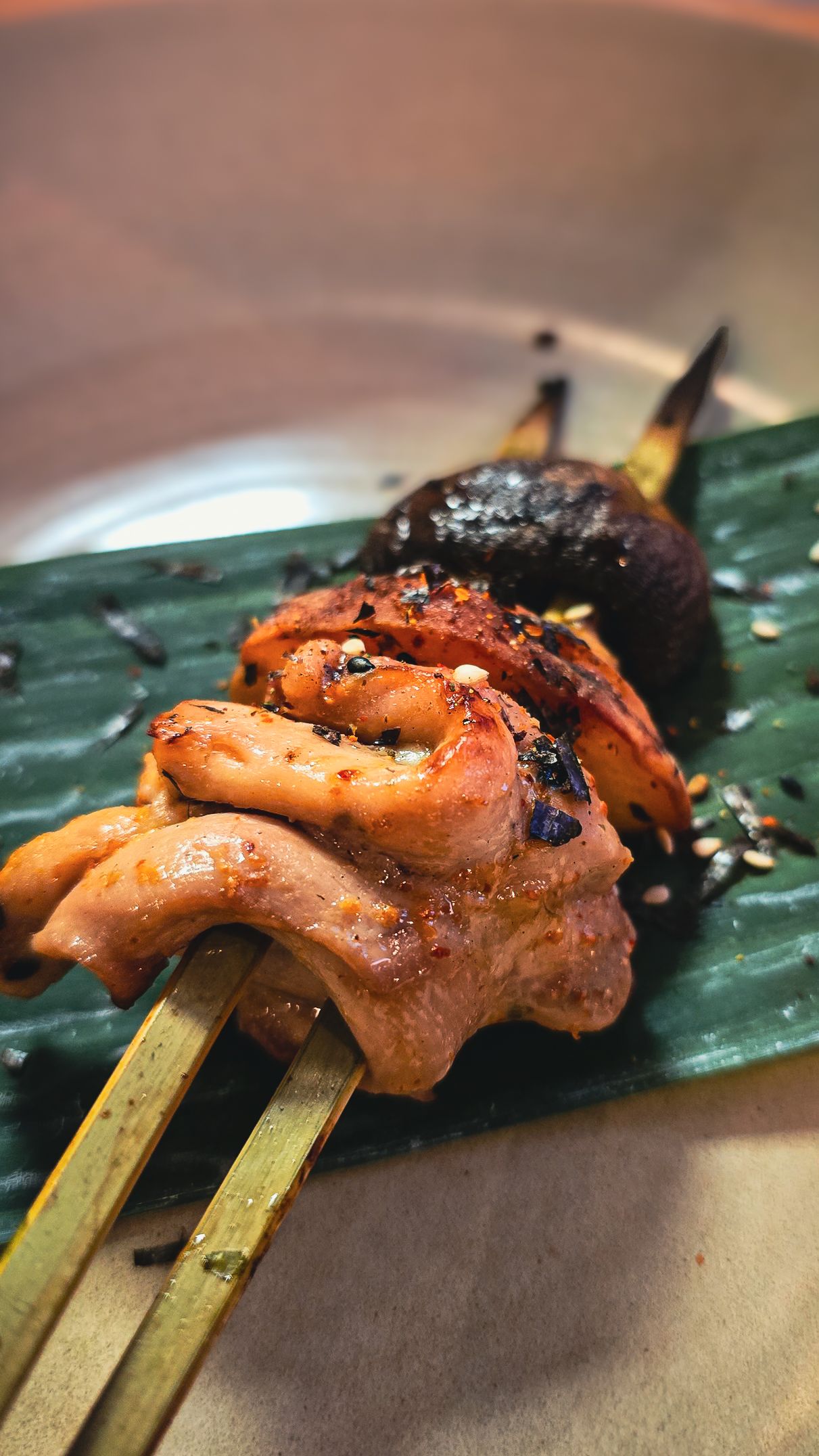 Grilled meat skewers with mushrooms on bamboo sticks, served on a green banana leaf with sesame seeds.