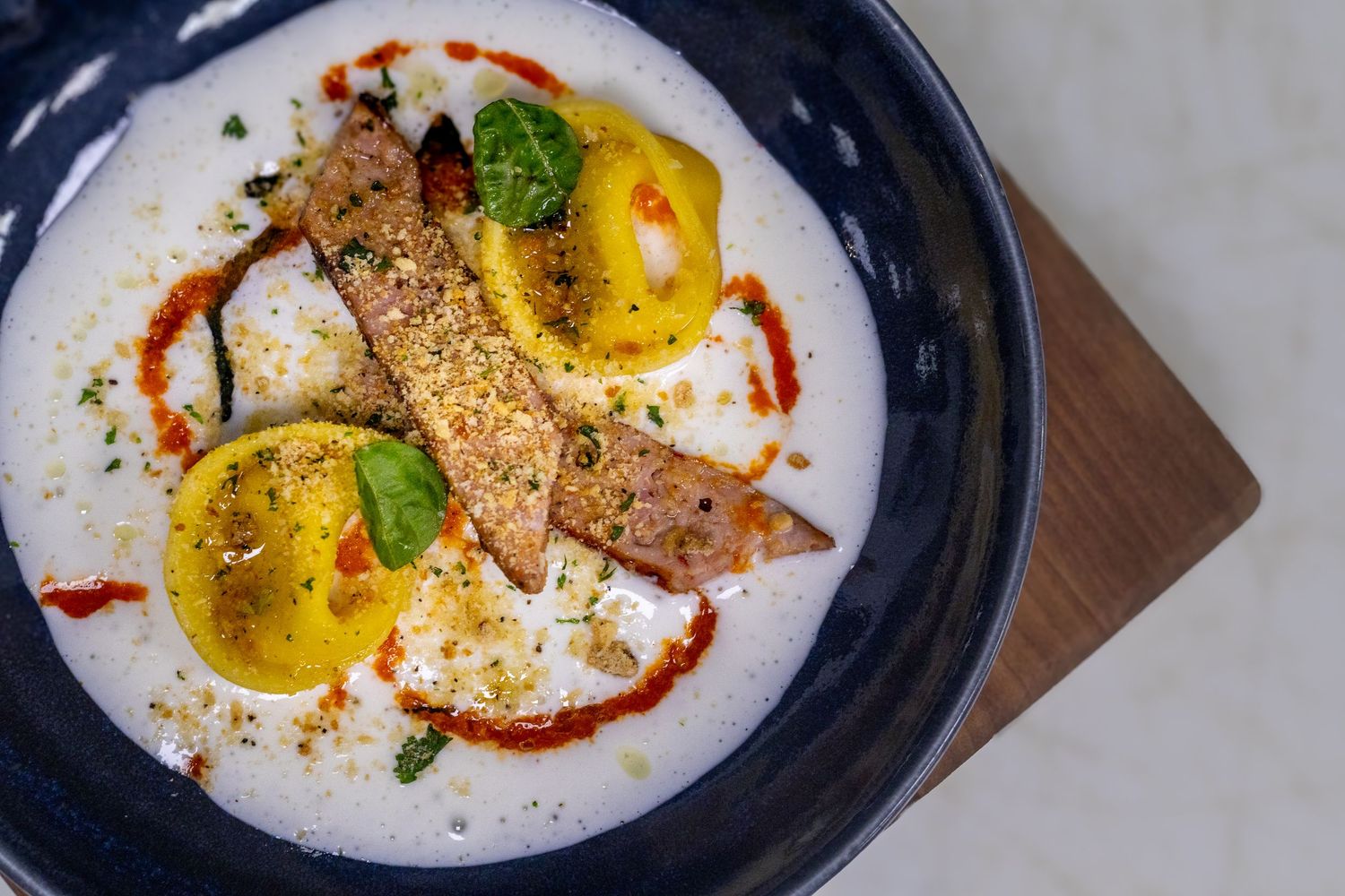 Elegant plated dish with seared fish, yellow tomatoes, and red sauce in creamy foam on a dark bowl.