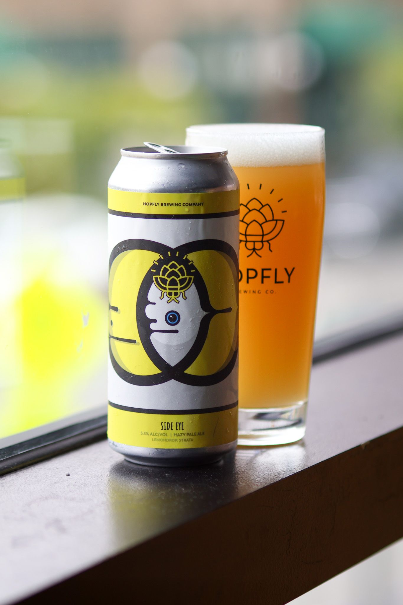 Hopfly Brewing Co. Side Eye IPA beer can beside a full pint glass on a bar ledge.