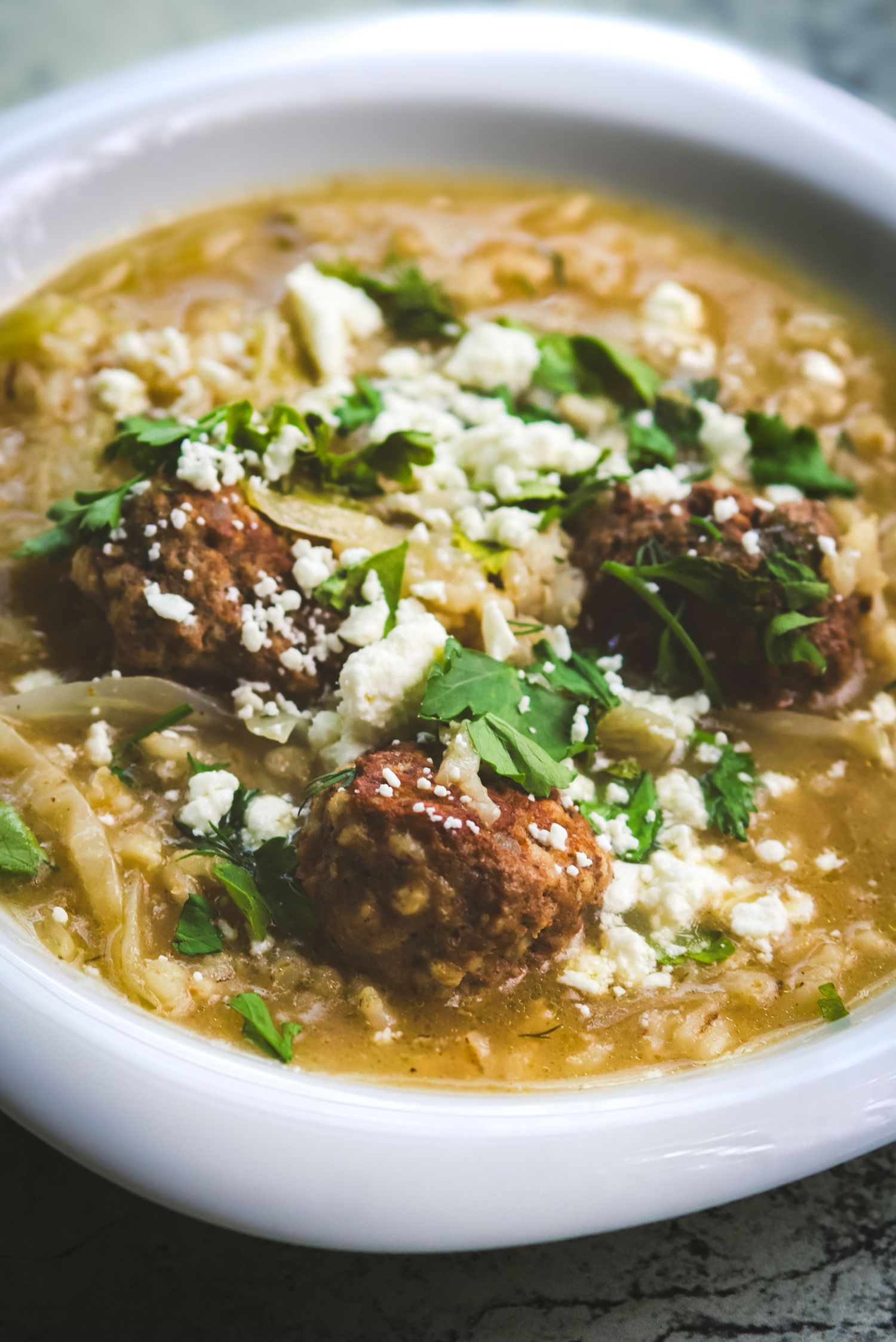 A rustic white bowl of meatball orzo soup garnished with crumbled white feta cheese and fresh chopped herbs.