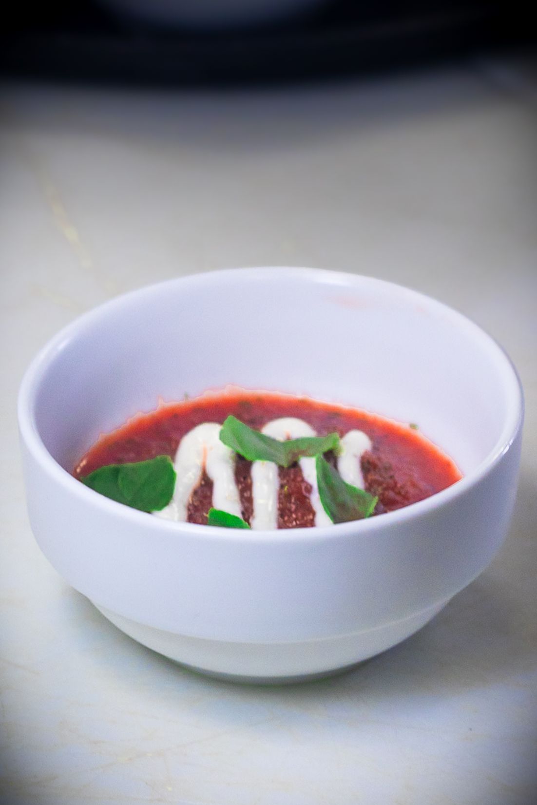 White bowl of tomato soup garnished with cream drizzle and fresh basil leaves on a light surface.