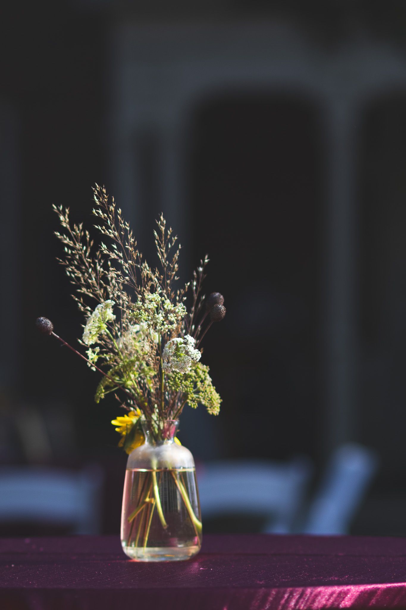 Wildflower bouquet in glass jar vase on purple velvet table with dark bokeh background.