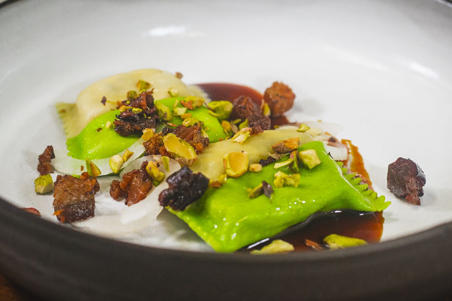 Gourmet dish with green ravioli, caramelized nuts, pistachios, and rich brown sauce in a white bowl.