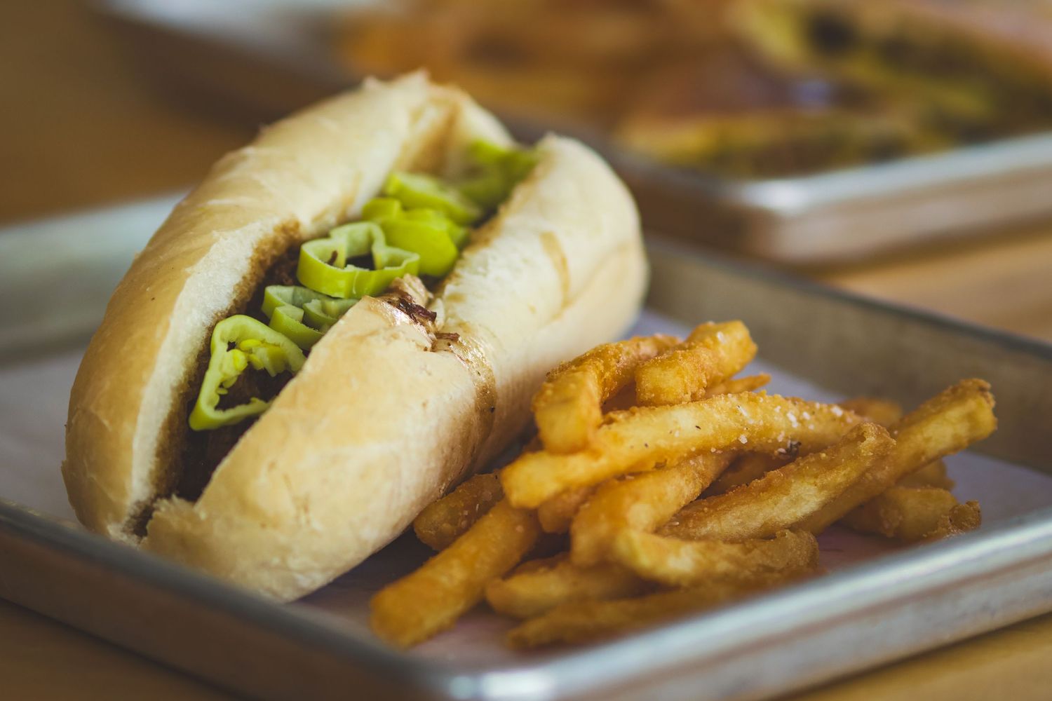 Savory hot dog with green peppers on a bun served with crispy seasoned fries on metal tray.