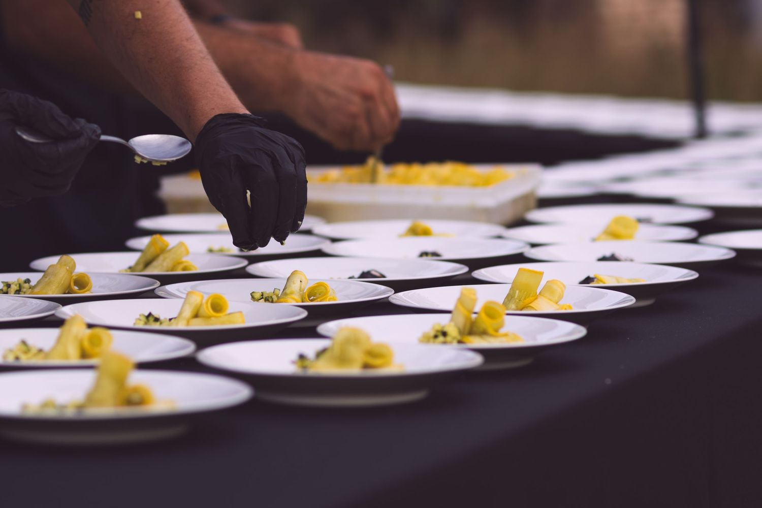 Chef in black gloves plating yellow pasta portions into rows of white bowls for service.