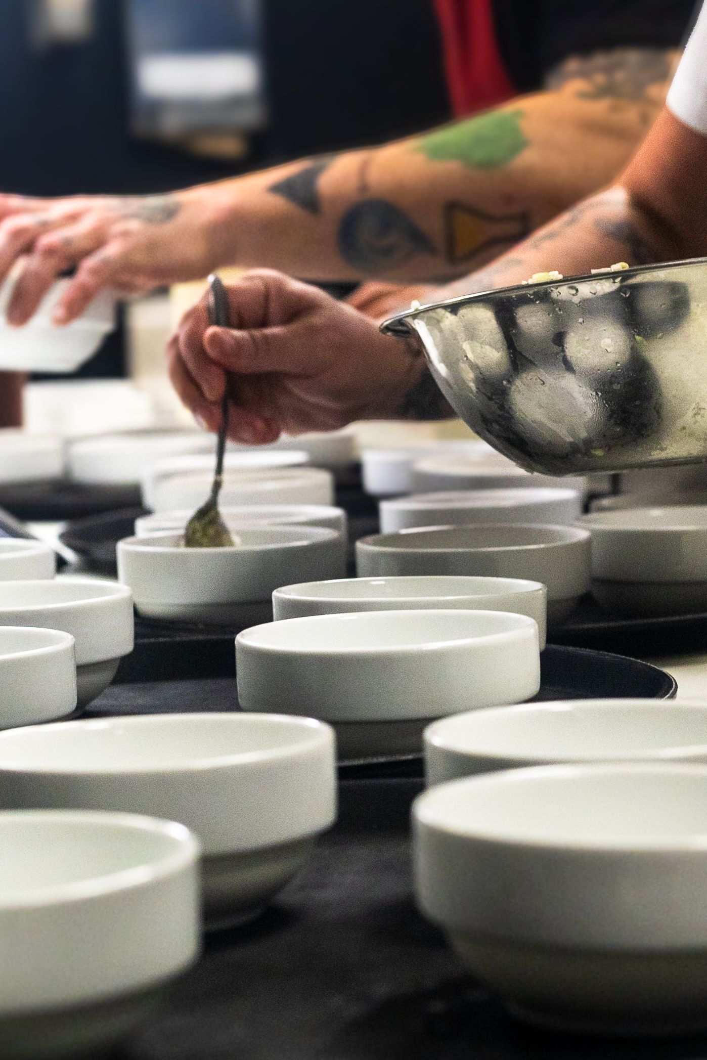 Tattooed chef carefully spooning sauce into rows of white ceramic bowls during professional kitchen prep service.